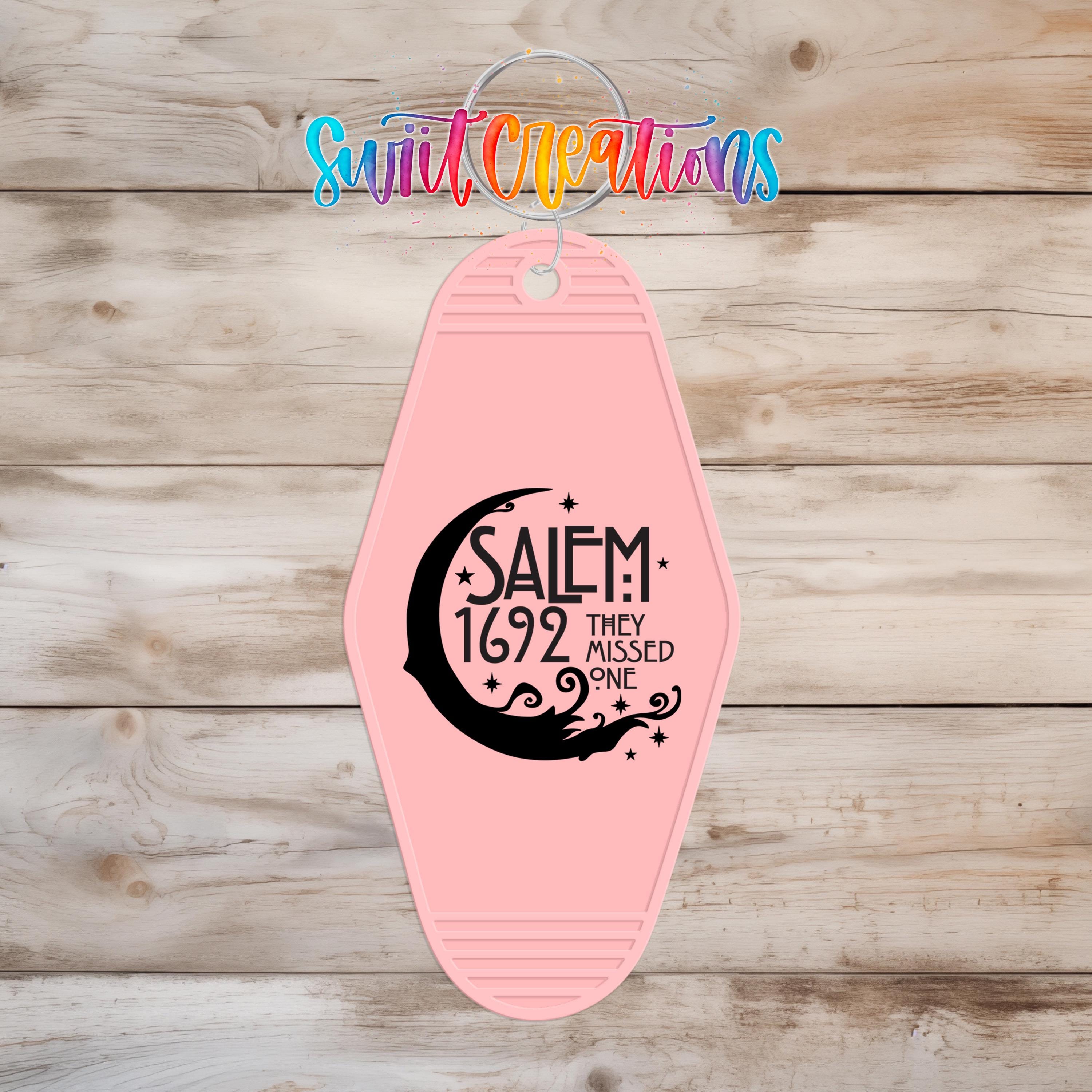 A pink keychain with a black design and text on it, hanging on a wooden surface.