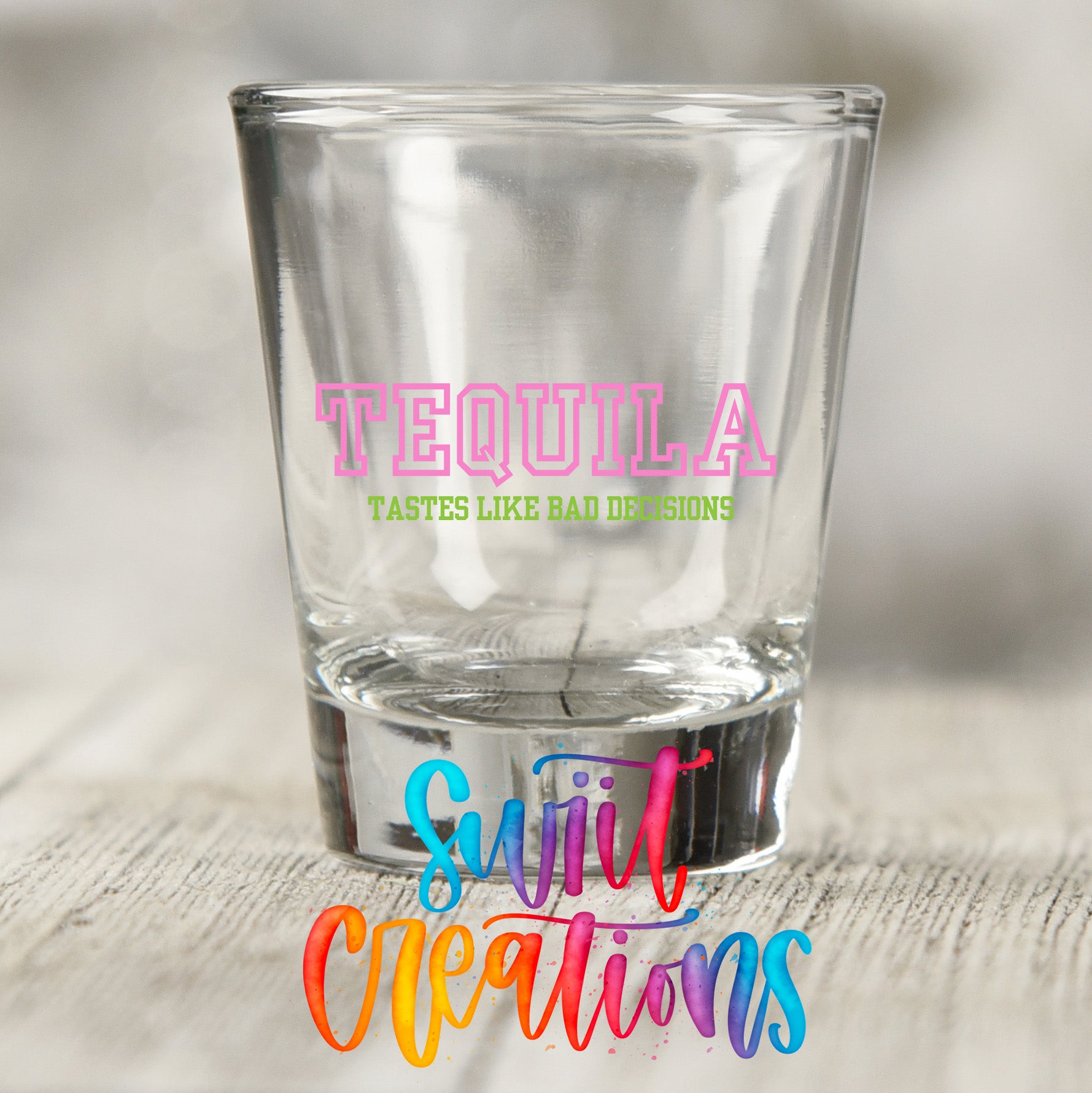 A clear shot glass with the word "TEQUILA" printed on it, along with the text "TASTES LIKE BAD DECISIONS" in pink and green. Below the shot glass, there is a colorful text logo that says "Sujiv Creations".
