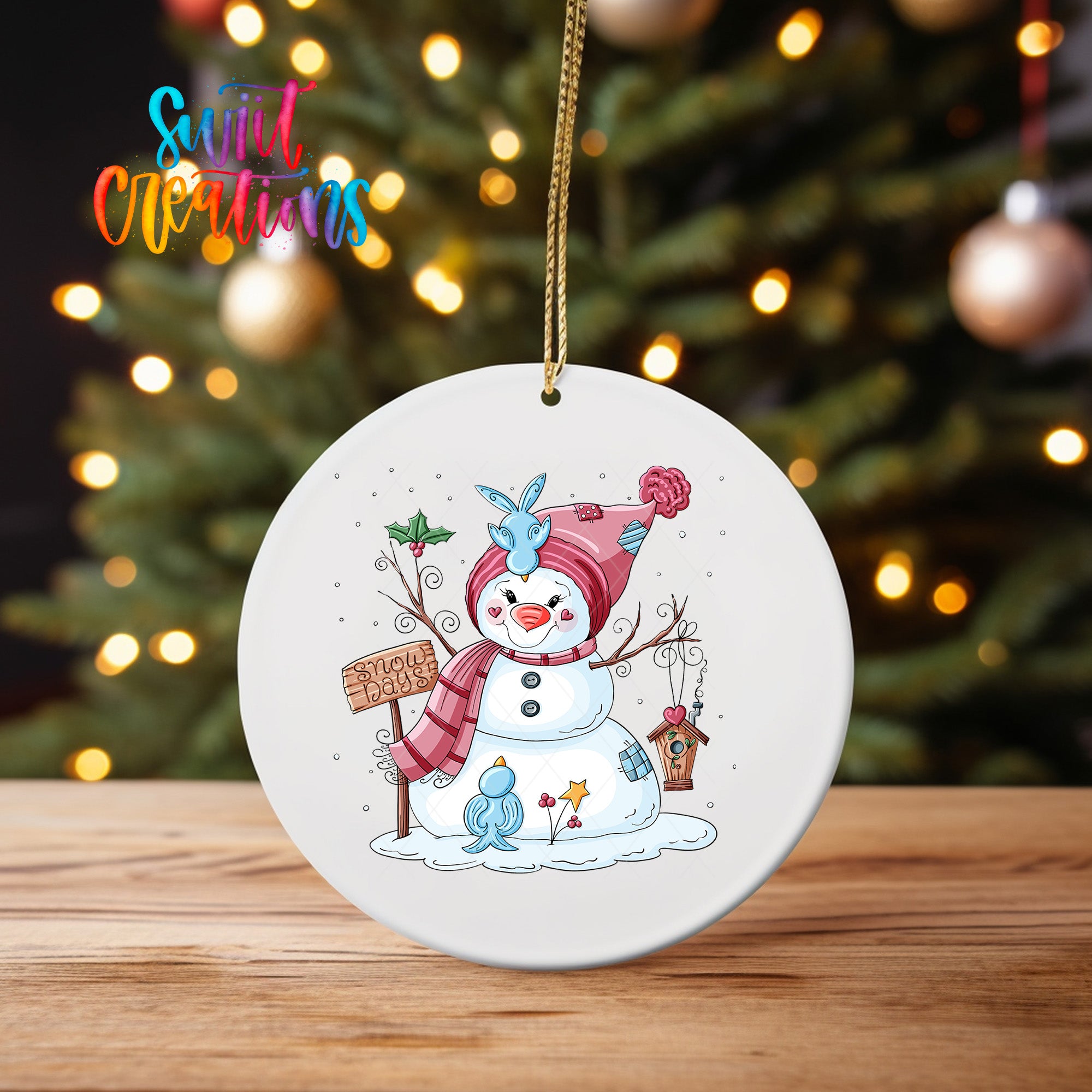 A white ornament featuring a snowman with a sign that says "Snowy Sign" is hanging on a wooden surface, with a blurred background of a Christmas tree and twinkling lights.