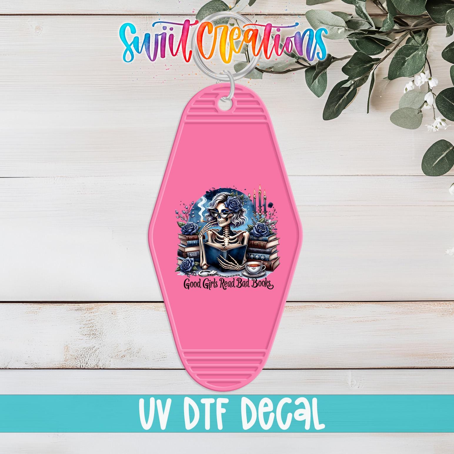A pink keychain with a skull design and the text "Good Girls Bad Books" printed on it, placed on a wooden surface with some greenery in the background.