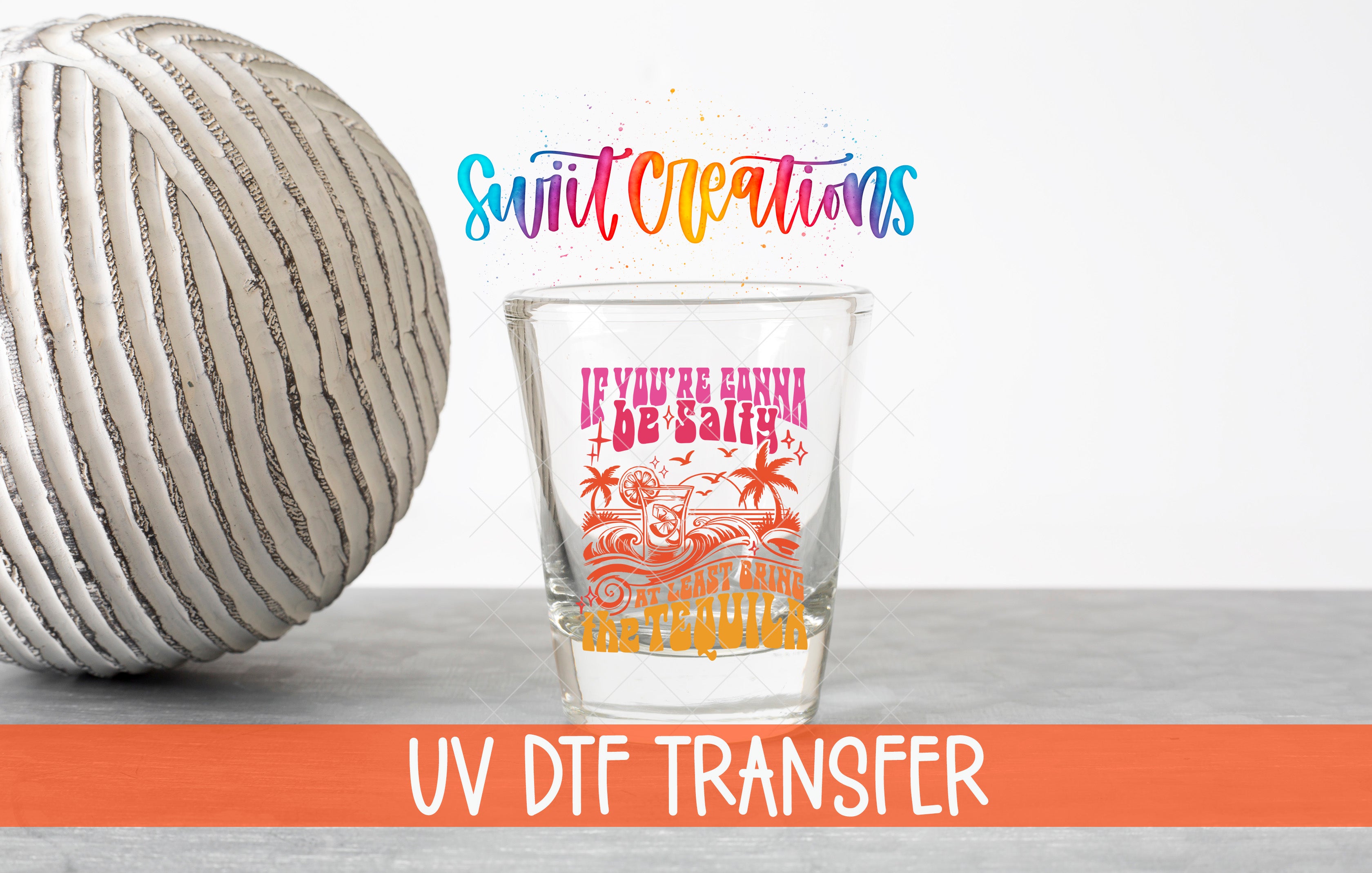 a glass with a colorful design and text, placed next to a white ball on a gray surface.
