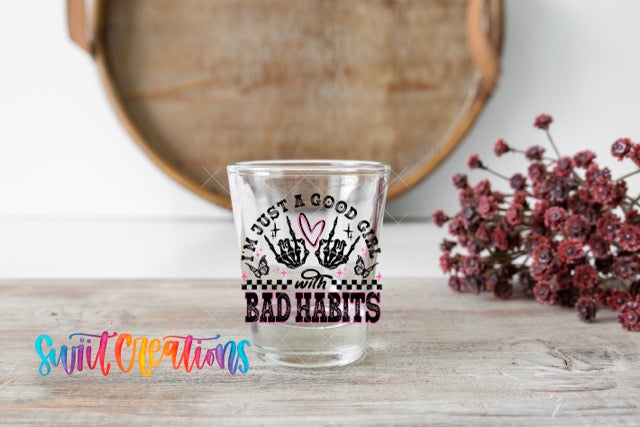 A clear shot glass with a hand-drawn design and text, placed on a wooden surface next to a bouquet of red flowers.