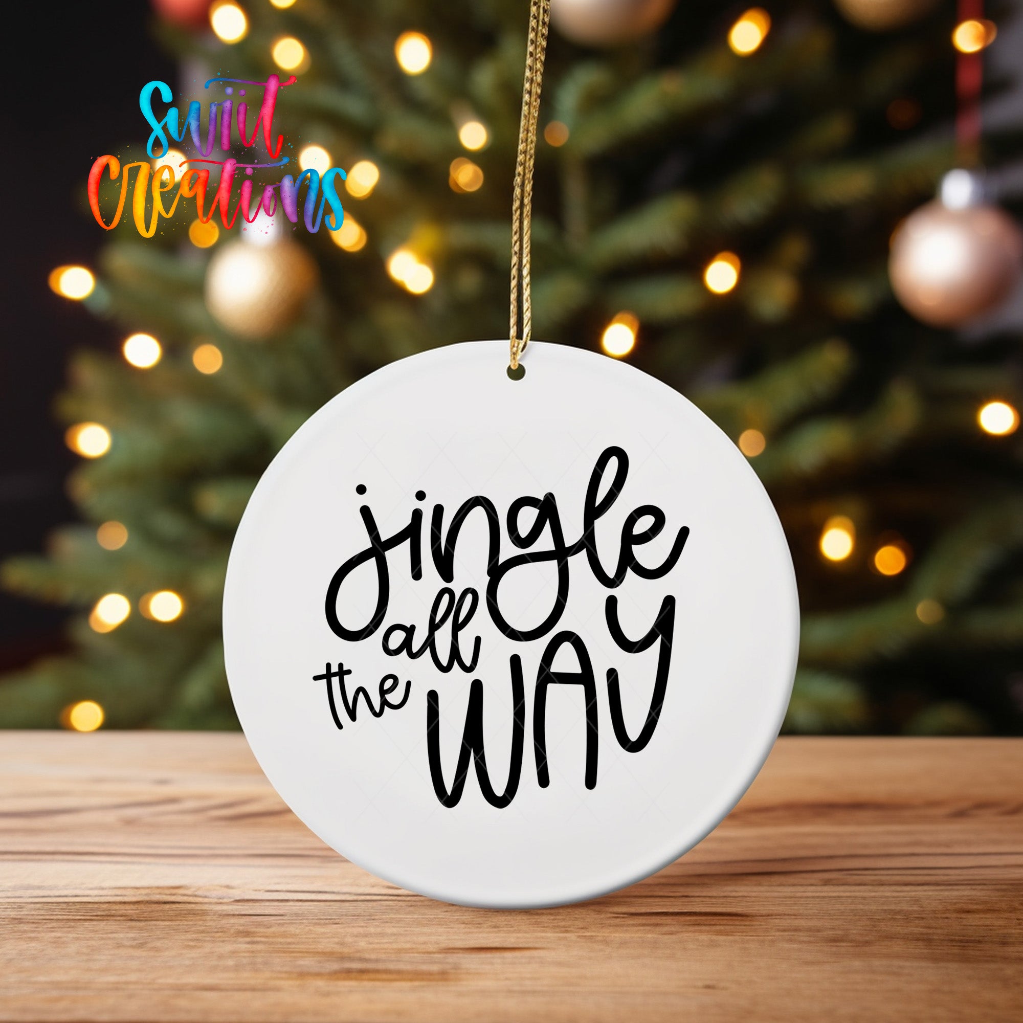 A white ornament with the text "Jingle all the way" hanging from a string, placed in front of a blurred Christmas tree with colorful lights.