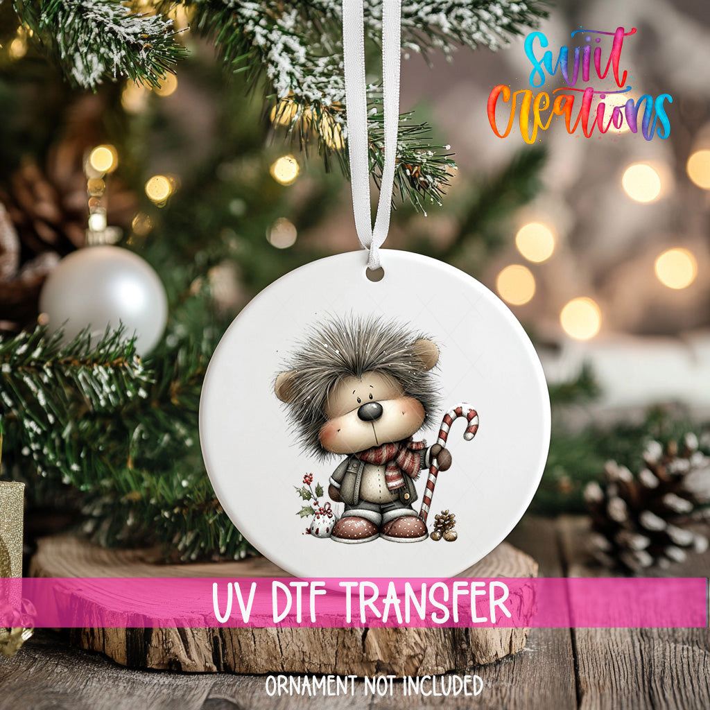 A white ornament with a cartoon hedgehog character is hanging on a Christmas tree, with a blurred background of pine branches and twinkling lights.