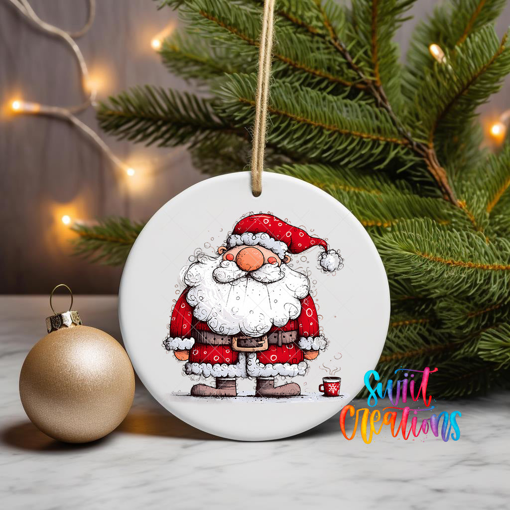 A white ceramic ornament featuring a cartoon illustration of Santa Claus wearing a red suit and hat, with a cup of coffee in front of him, is displayed on a table next to a gold ornament and a Christmas tree.