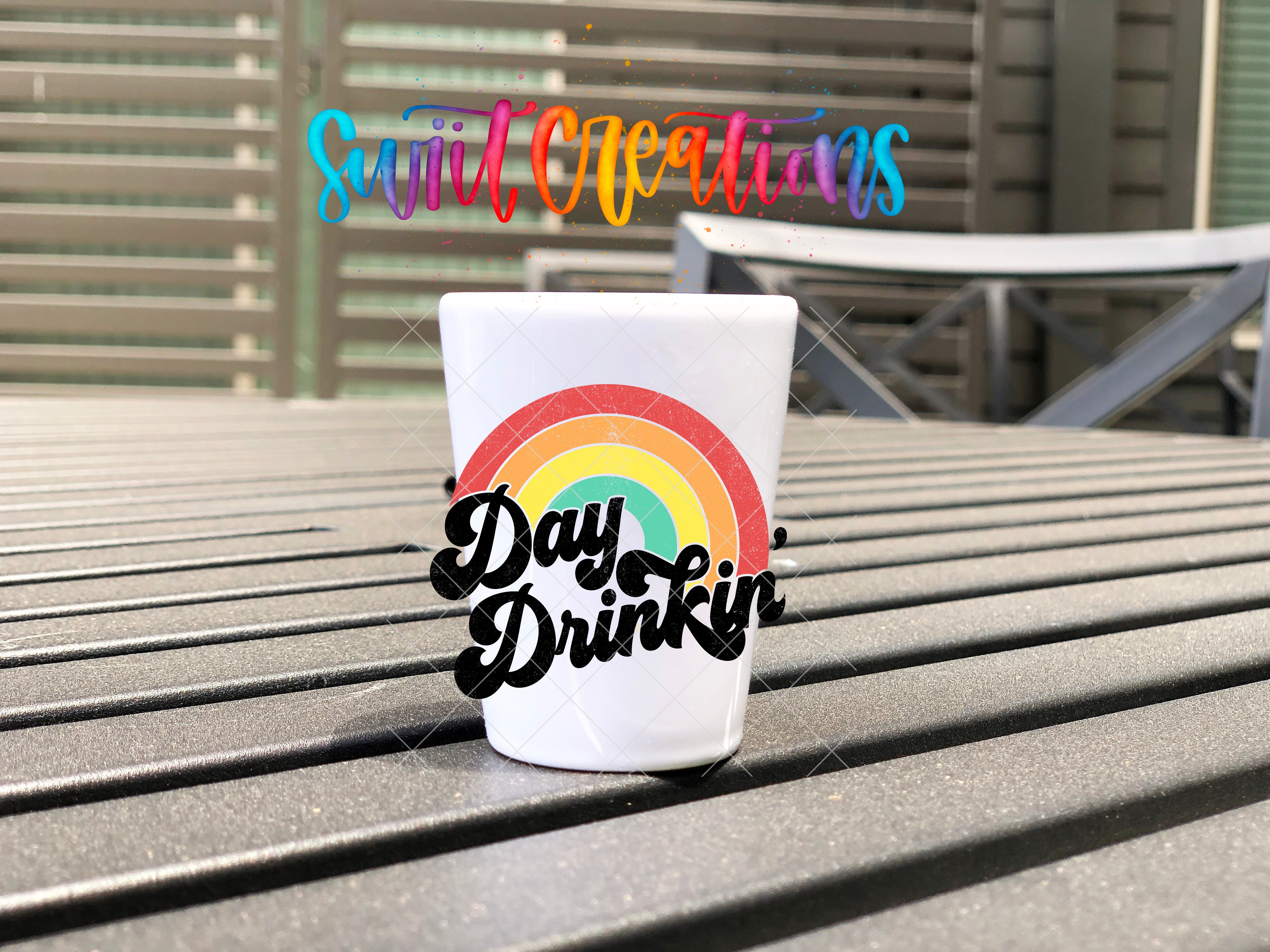 A white cup with a rainbow design and the text "Day Drinkin" on it, placed on a wooden surface.