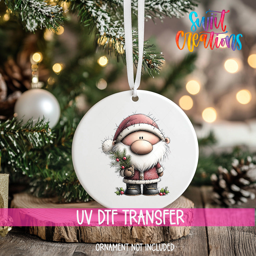 A white ornament with a cartoon Santa Claus figure is hanging on a Christmas tree, with a blurred background of pine branches and twinkling lights.