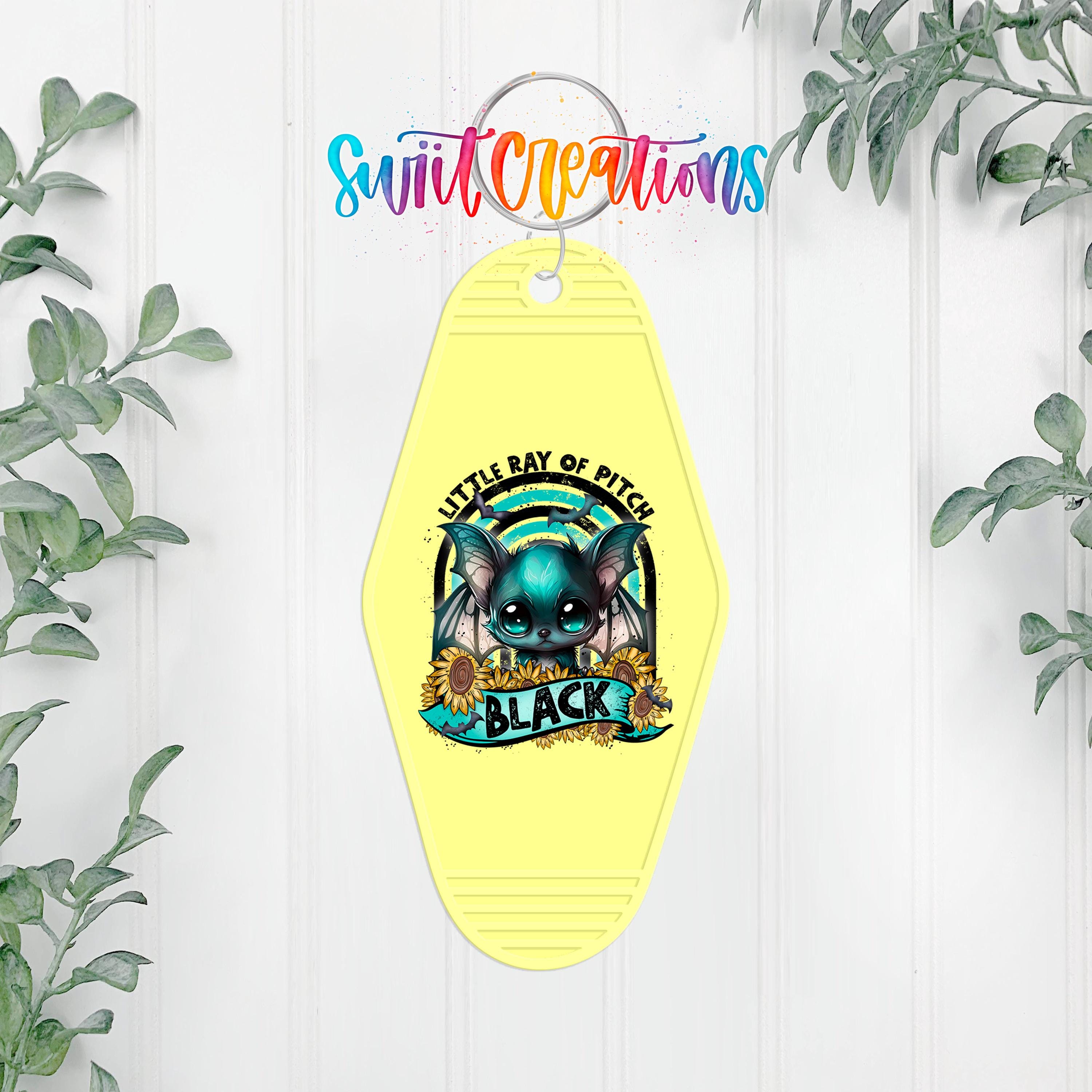 A yellow keychain with a bat and sunflower design, hanging on a white wall with green plants.