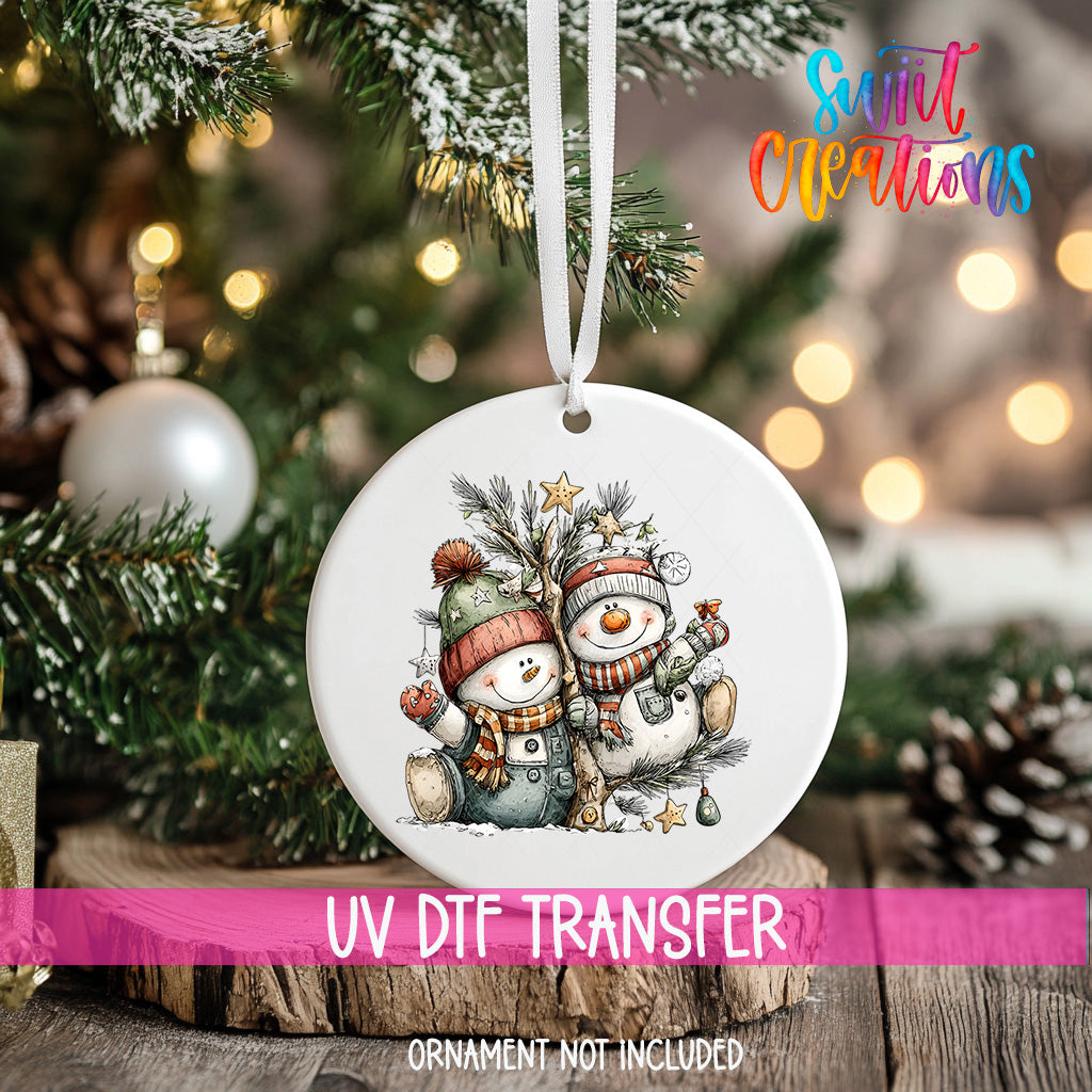 A white ornament with a snowman and star on a Christmas tree, with a pink ribbon around it.
