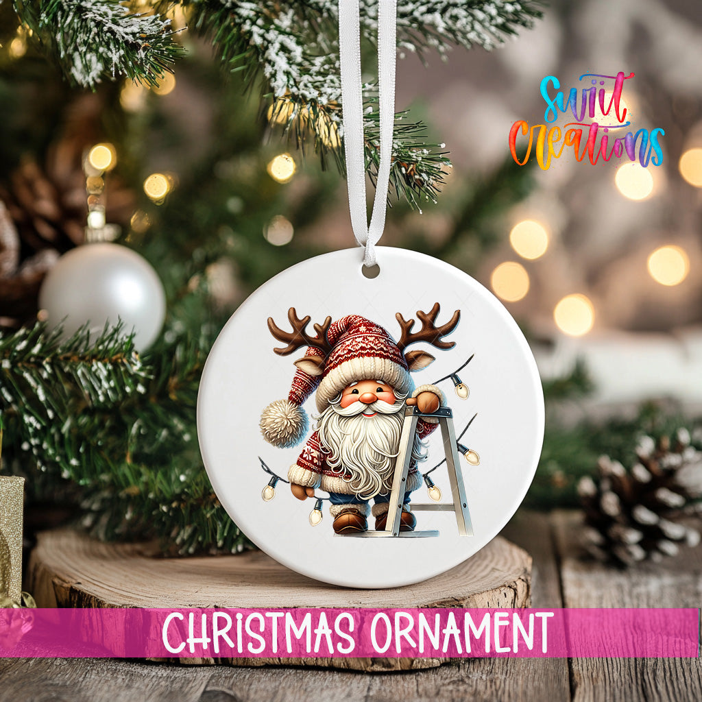 A white Christmas ornament featuring a smiling Santa Claus with antlers and a ladder, hanging on a tree branch with a blurred background of lights.