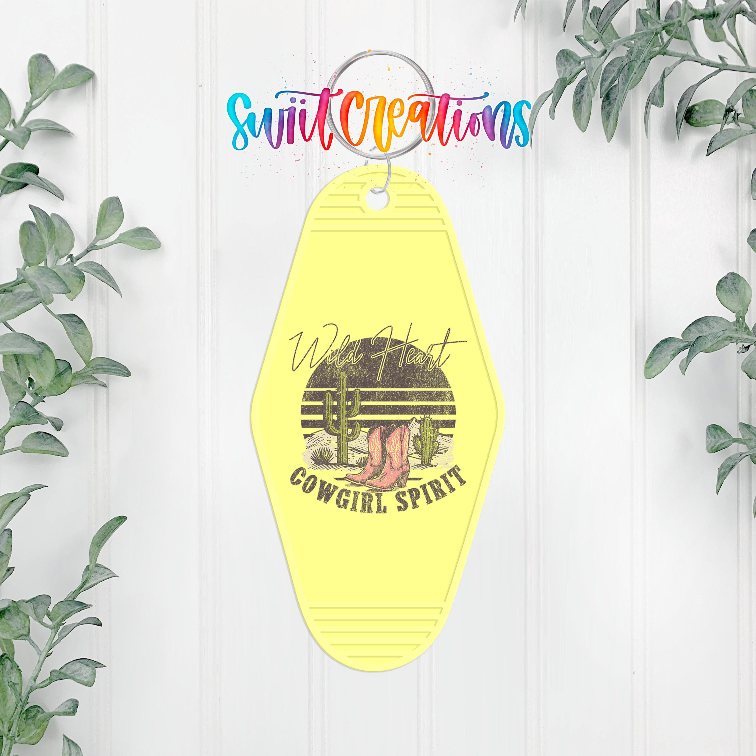 A yellow keychain with a graphic design featuring a cactus and the text "Wild Heart Cowgirl Spirit" is hanging on a white wall.