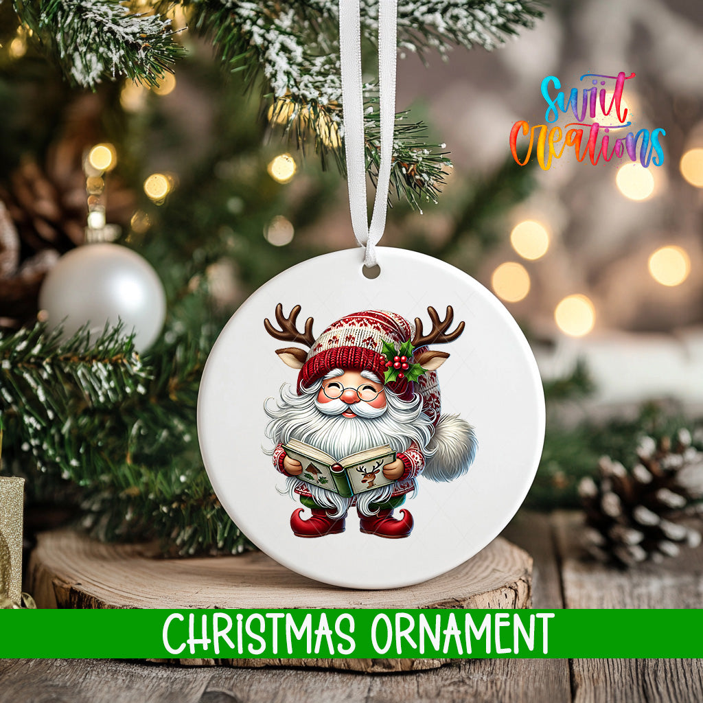 A white Christmas ornament featuring a smiling Santa Claus with antlers and a book, hanging on a tree branch with a blurred background of lights and pine cones.