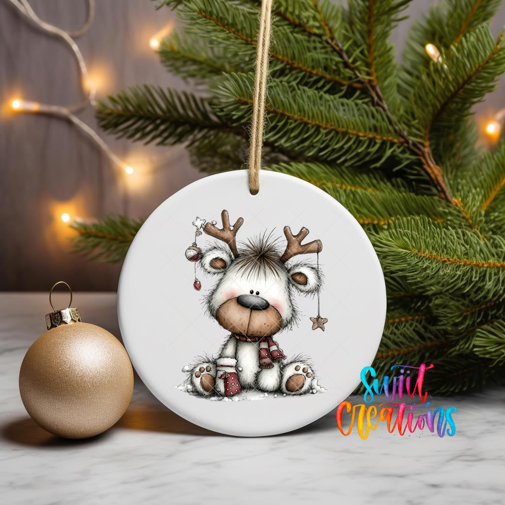 A white ceramic ornament featuring a cartoon reindeer with antlers and a star, hanging on a string next to a gold ornament and a Christmas tree.