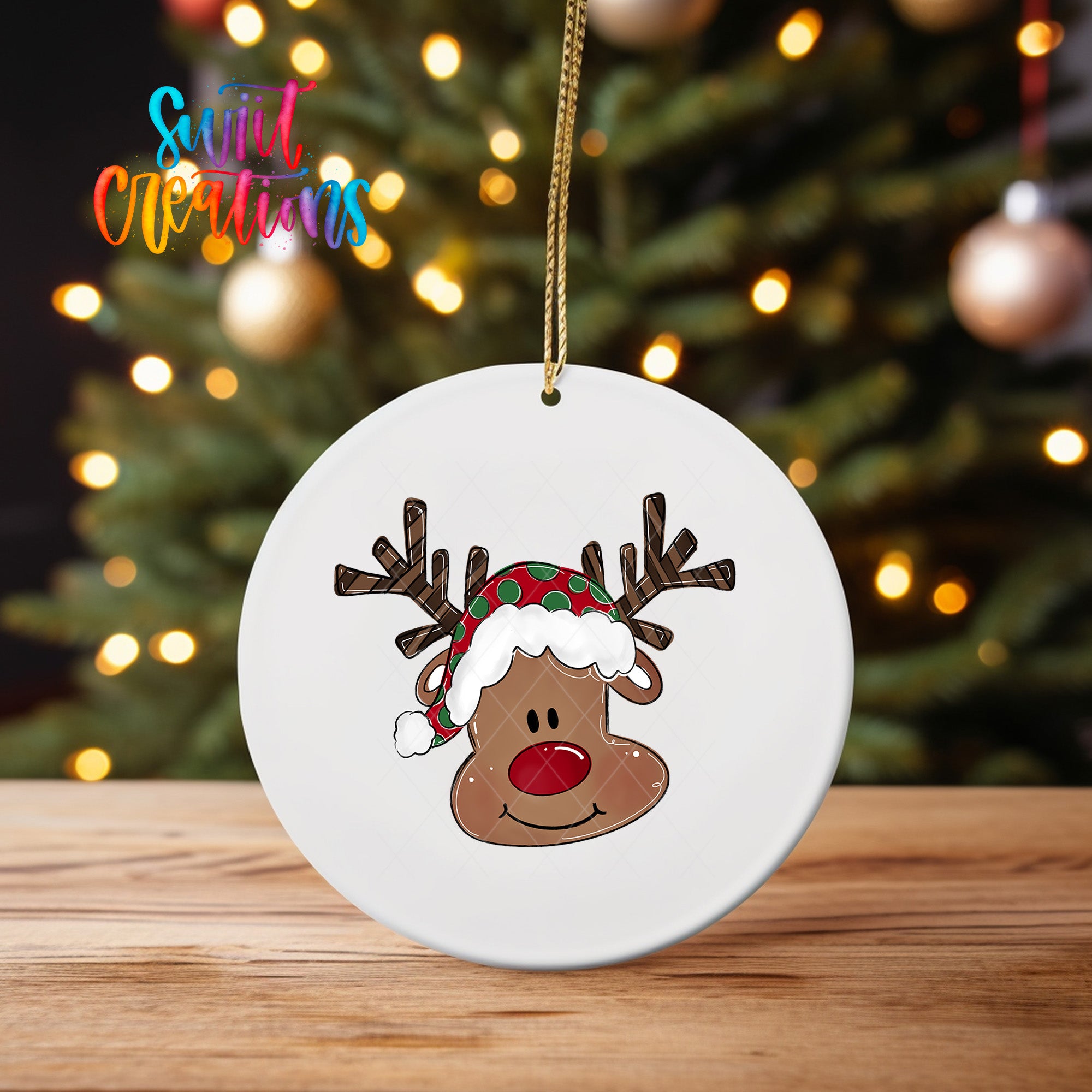 A white ornament with a cartoon reindeer wearing a Santa hat is displayed on a wooden surface, with a blurred background of a Christmas tree and colorful lights.