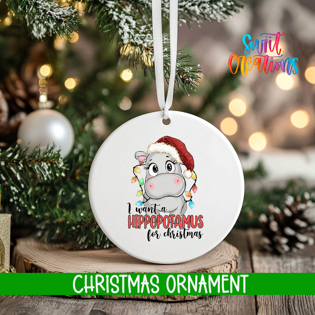 A white Christmas ornament with a cartoon image of a hippopotamus wearing a Santa hat and the text "I want a hippopotamus for Christmas" is hanging on a tree.