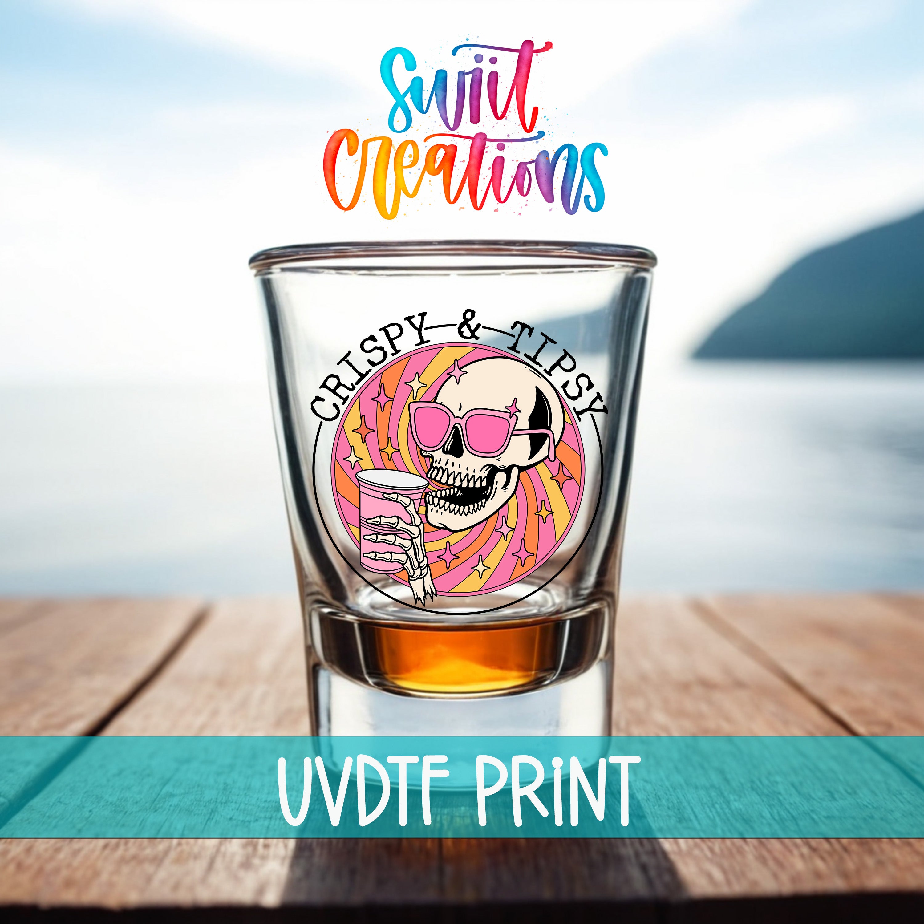 A shot glass with a skull design and the text "CRISPY & TIPSY" printed on it, placed on a wooden surface with a body of water in the background.