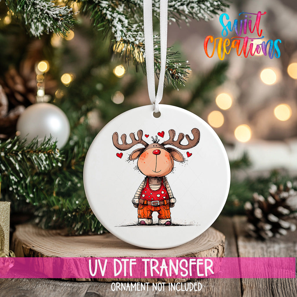 A white ceramic ornament with a cartoon reindeer design hangs on a Christmas tree, surrounded by pine branches and twinkling lights.
