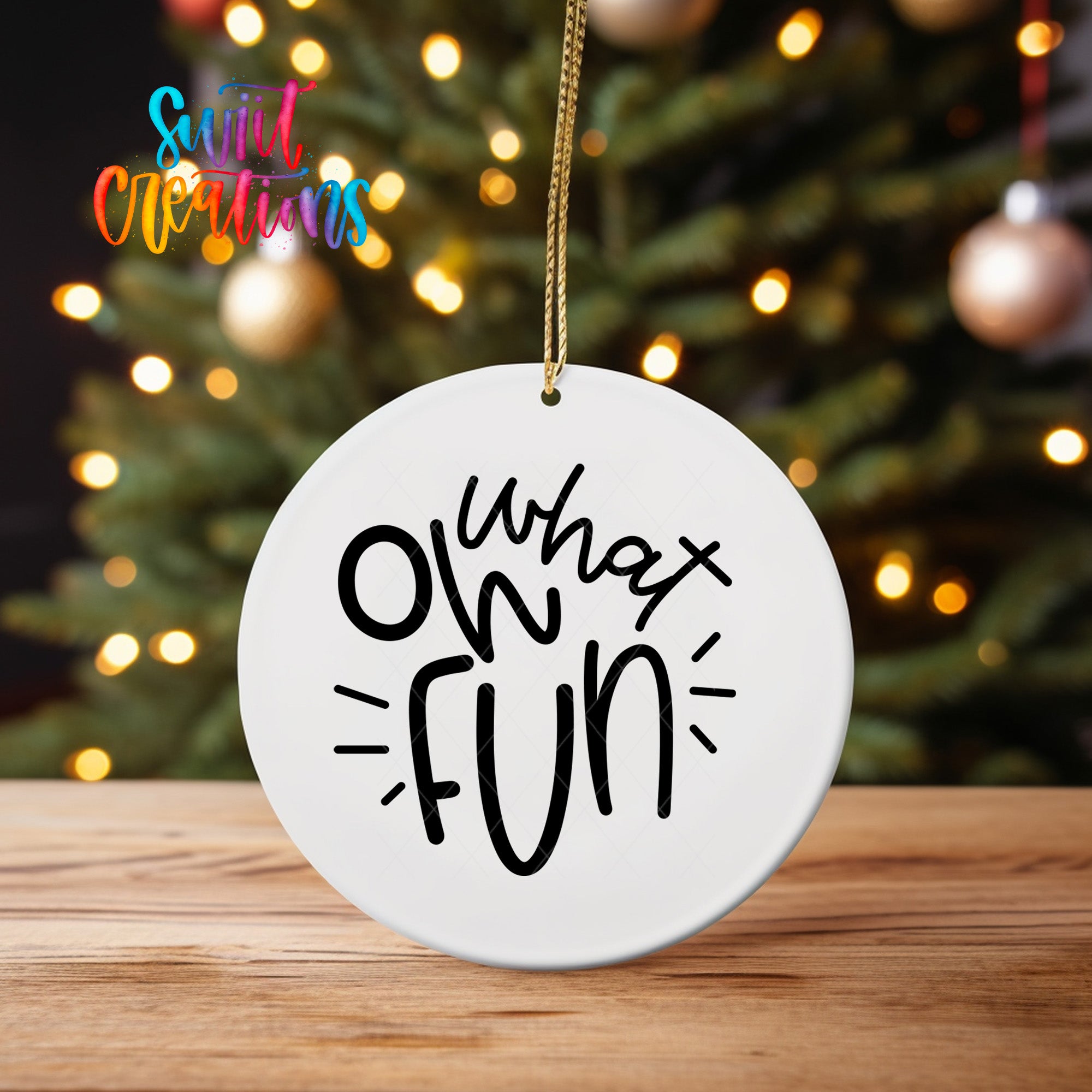A white ornament with the text "What fun" written on it, hanging from a string in front of a blurred Christmas tree with colorful lights.