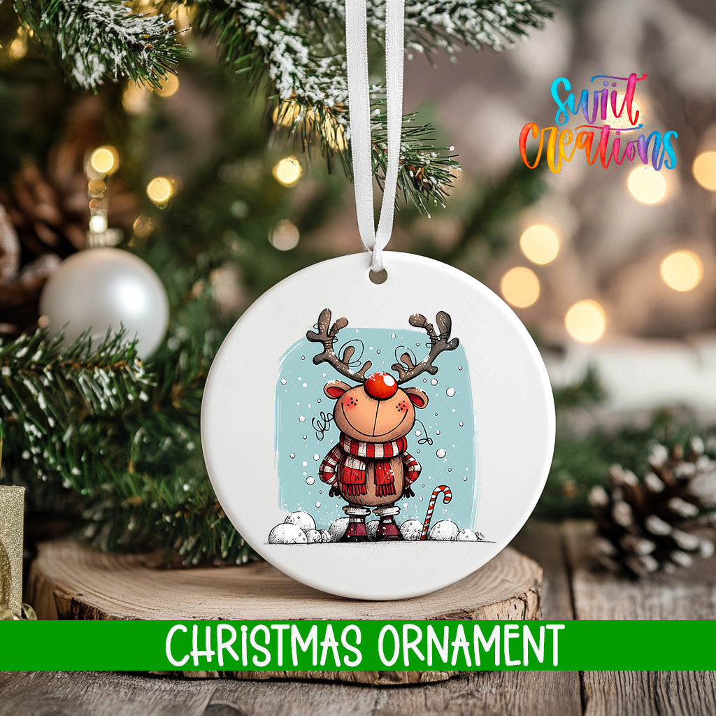A white Christmas ornament featuring a cartoon reindeer with antlers and a scarf, hanging on a tree branch with snowflakes falling around it.