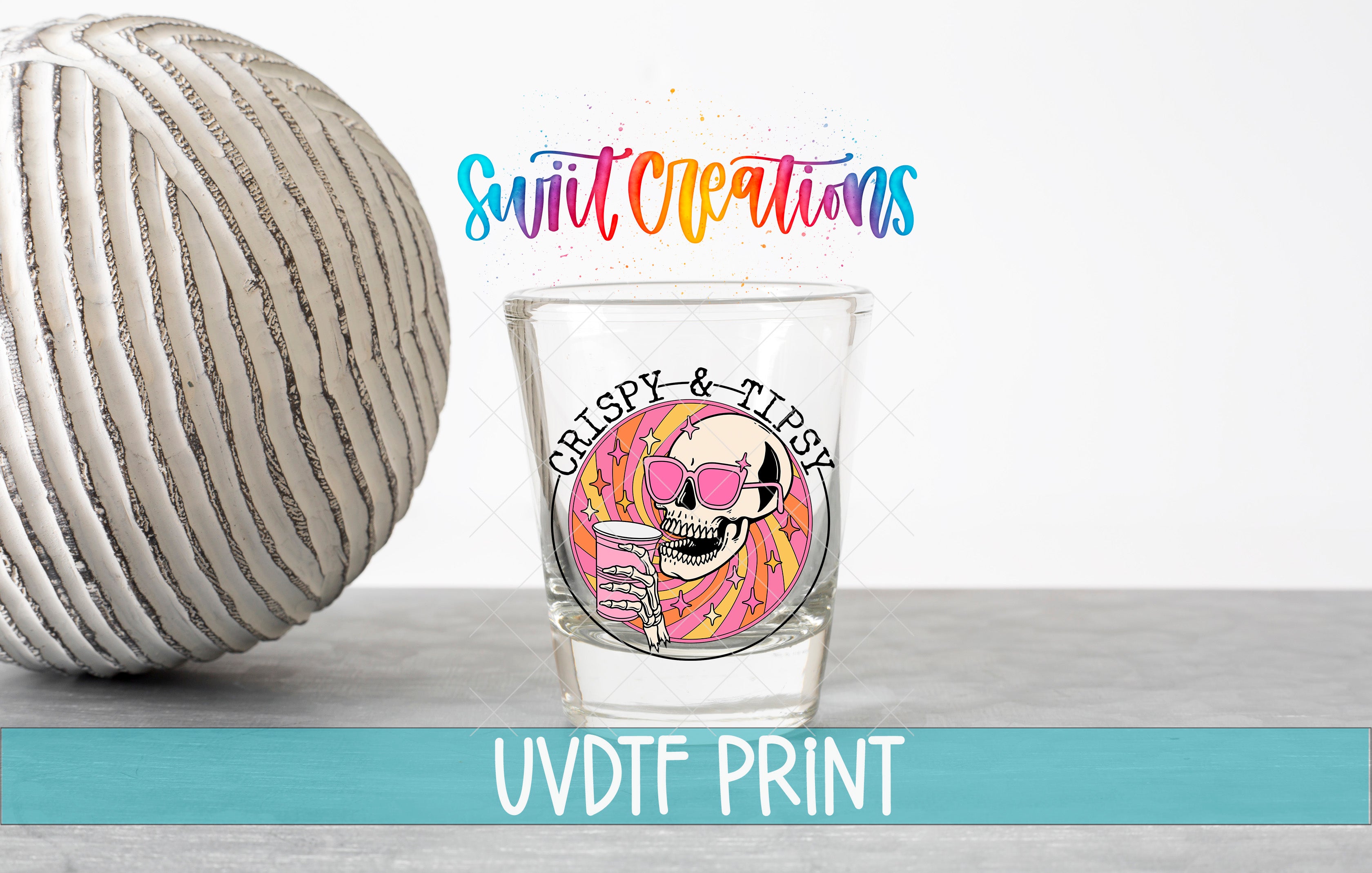 a shot glass with a skull wearing sunglasses and holding a drink, along with a striped ball and text that reads "Swift Creations" and "CRISPY & TISSY".