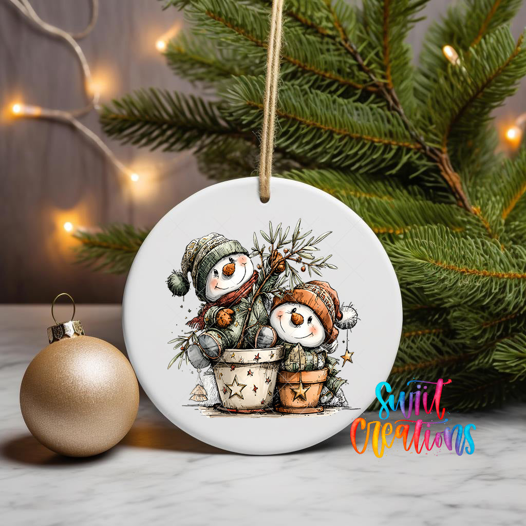 A white ceramic ornament featuring two snowmen sitting in a pot, surrounded by Christmas decorations, is displayed on a table with a gold ornament and a Christmas tree in the background.