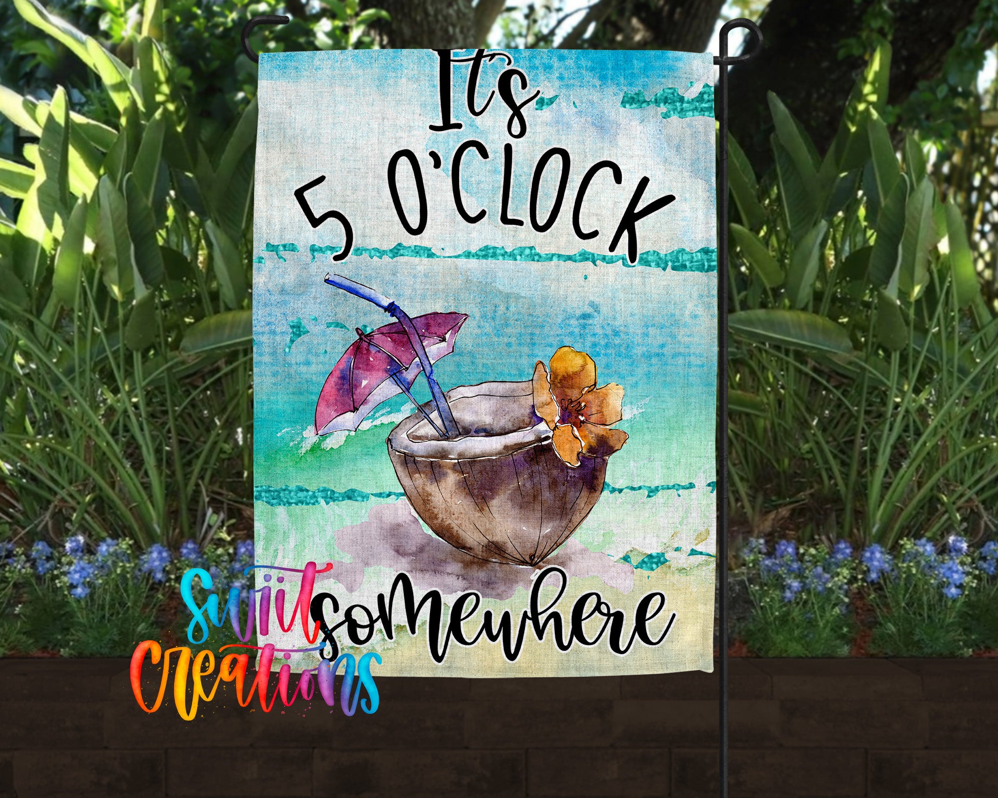 A colorful garden flag with a tropical drink and umbrella design, surrounded by lush greenery and flowers.