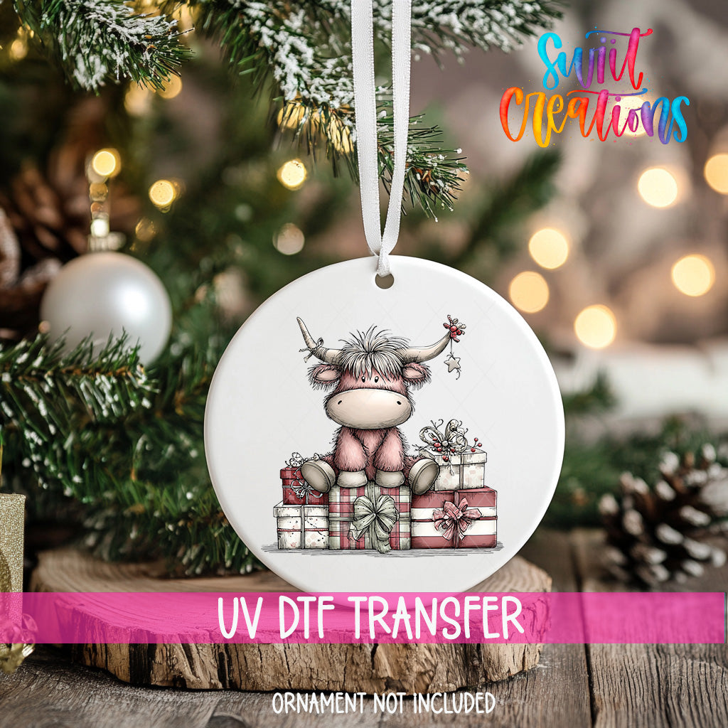 A white ornament with a cartoon cow sitting on a pile of presents is hanging on a Christmas tree.