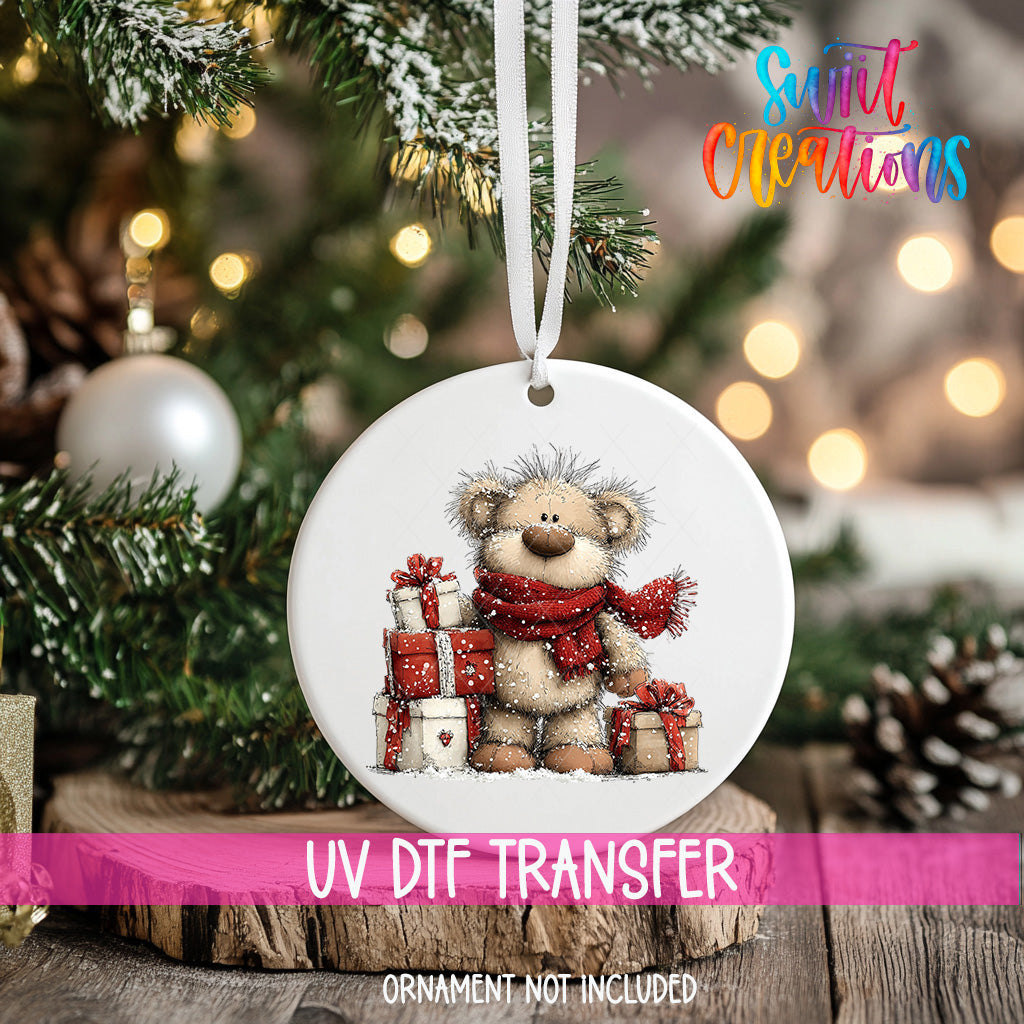 A white ornament with a teddy bear and presents is hanging on a Christmas tree, with a pink ribbon around it.
