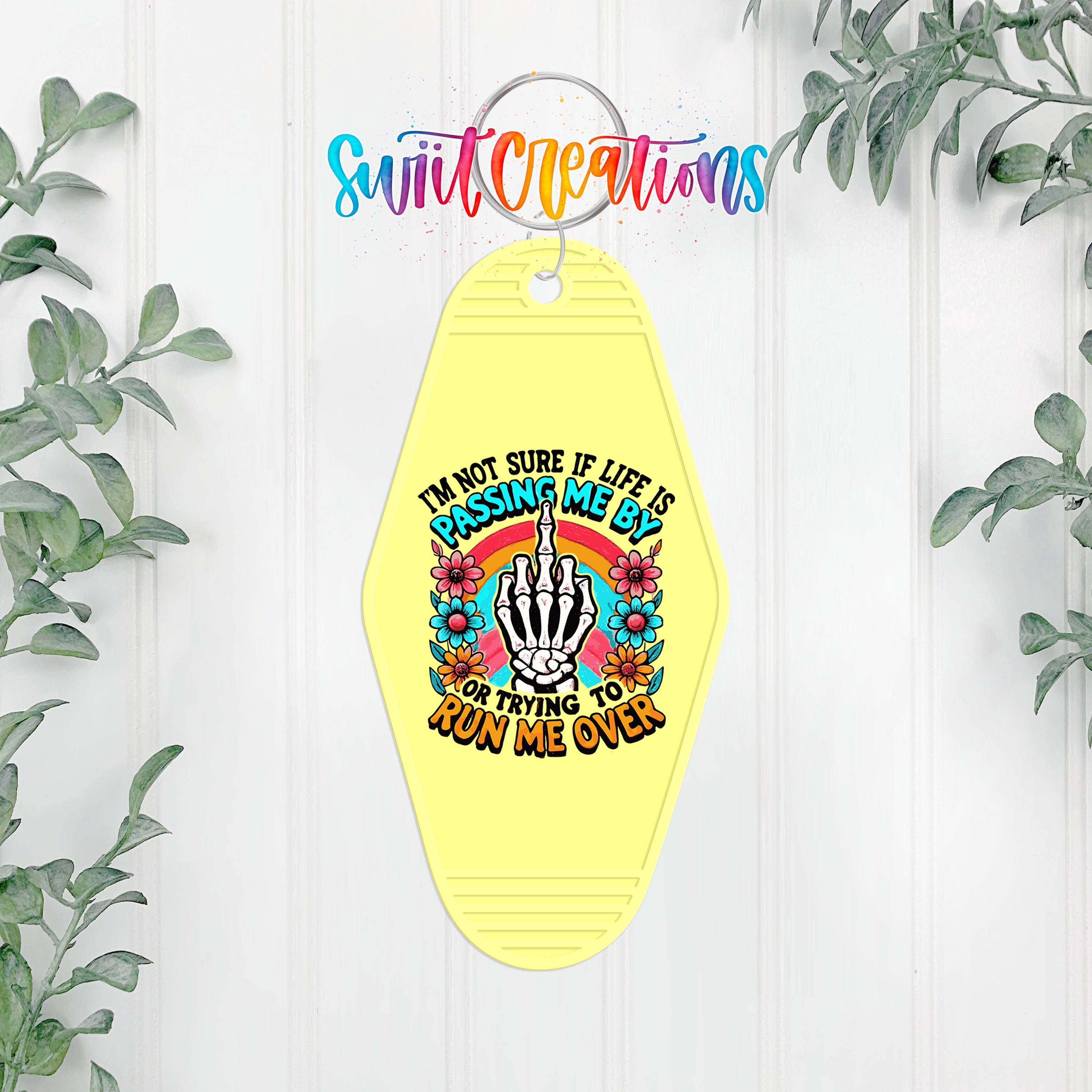A yellow keychain with a colorful graphic design and text, hanging on a white wall with greenery in the background.