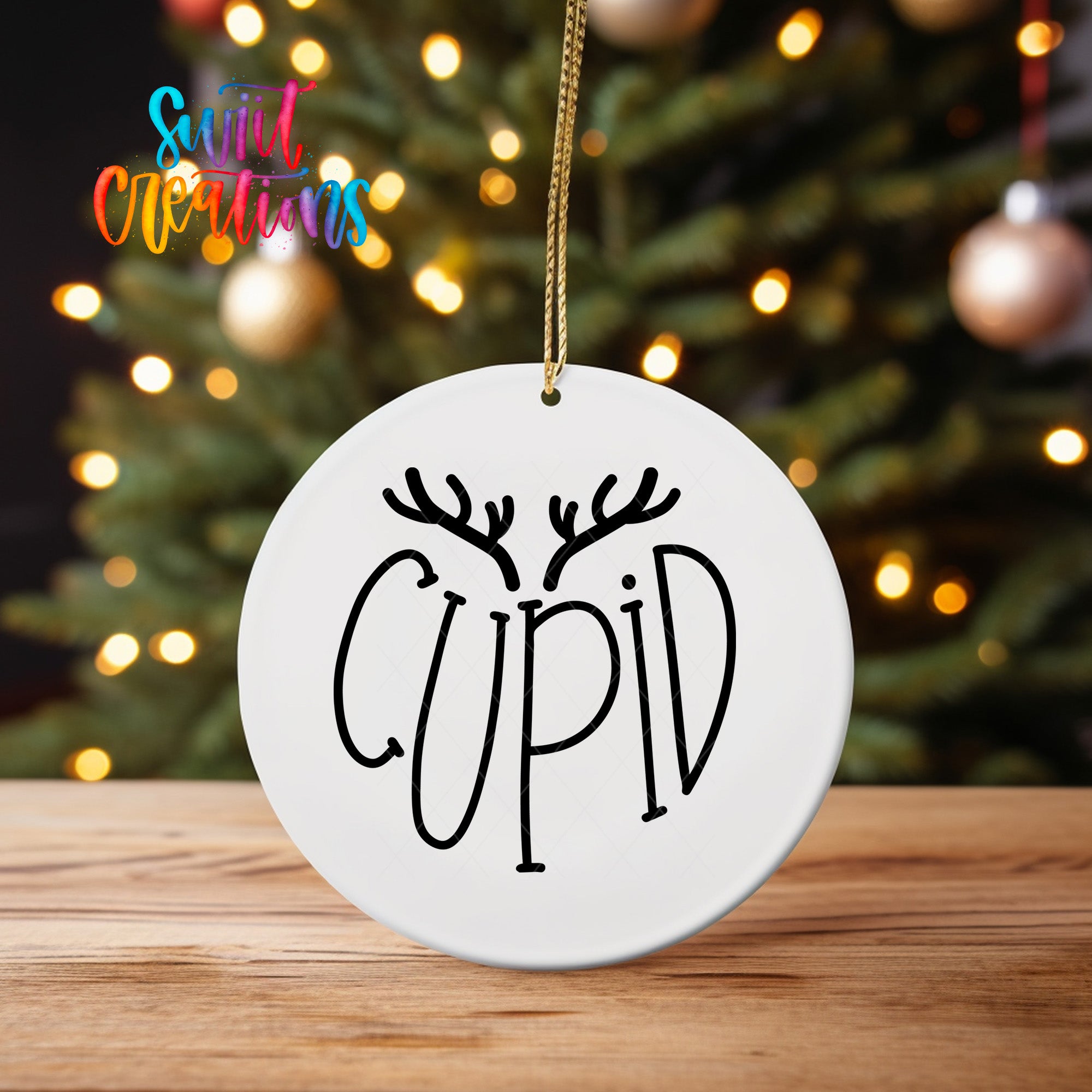 A white ceramic ornament with the word "CUPID" written on it, hanging from a string. The ornament is placed on a wooden surface, with a blurred background featuring a Christmas tree and colorful lights.