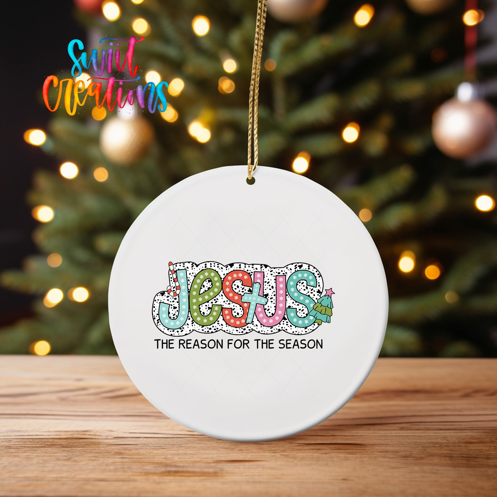 A white ceramic ornament with the word "JESUS" written on it, along with the text "THE REASON FOR THE SEASON" below it, is hanging from a string in front of a blurred Christmas tree.