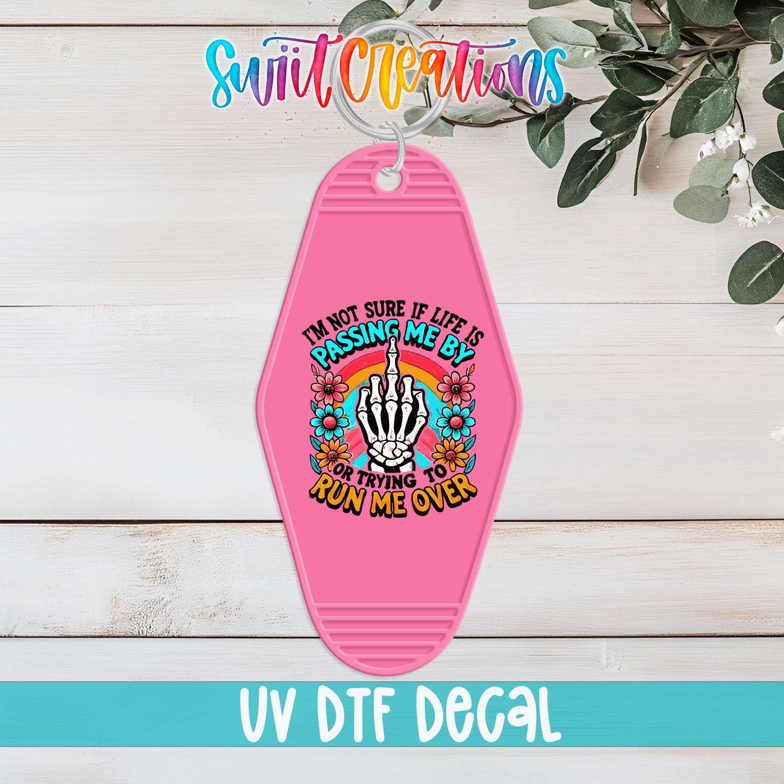 A pink keychain with a colorful graphic design and text, placed on a wooden surface with greenery in the background.