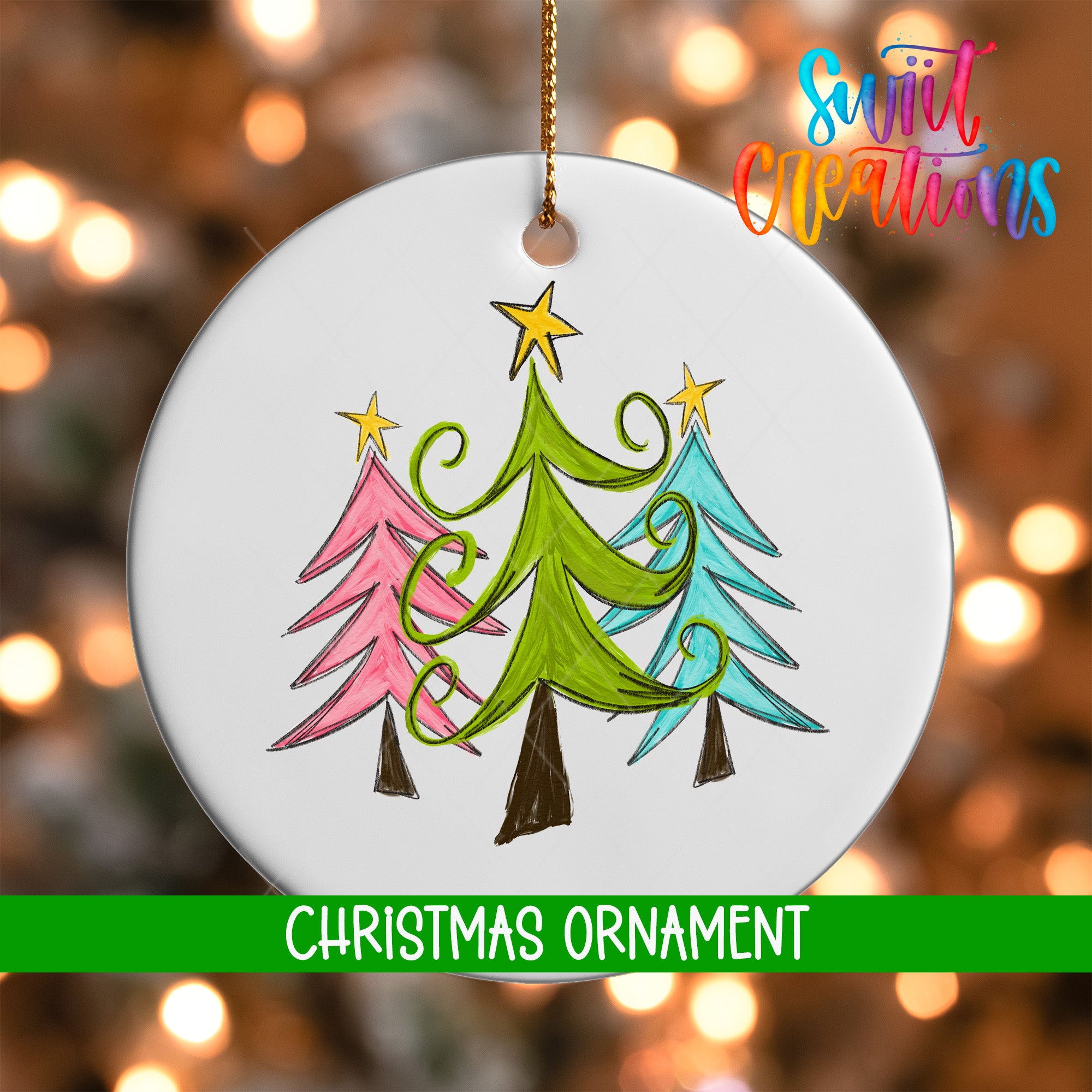 A white Christmas ornament with a drawing of three stylized Christmas trees in different colors, including pink, green, and blue, hanging against a blurred background with colorful lights.