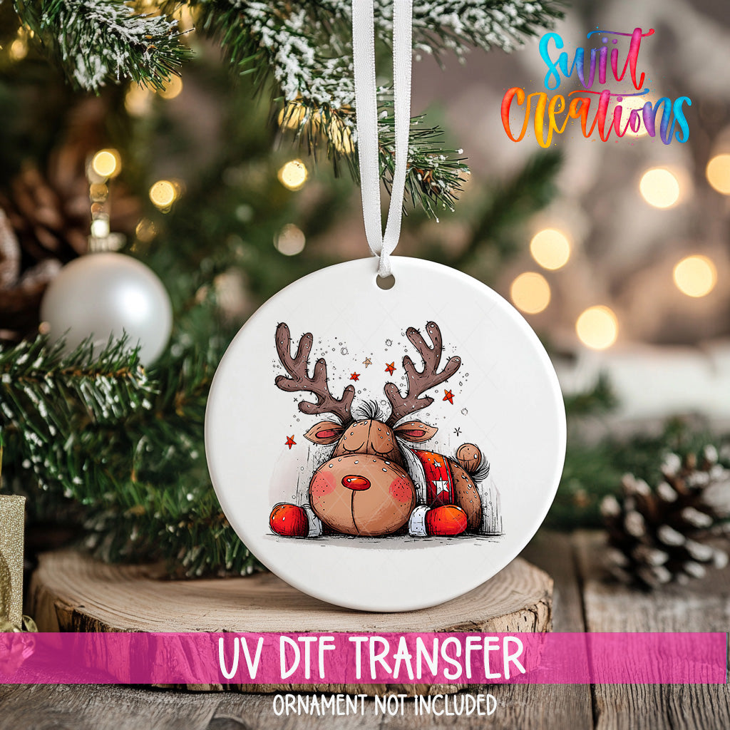 A white ceramic ornament with a cartoon reindeer design hangs on a Christmas tree, surrounded by pine branches and twinkling lights.