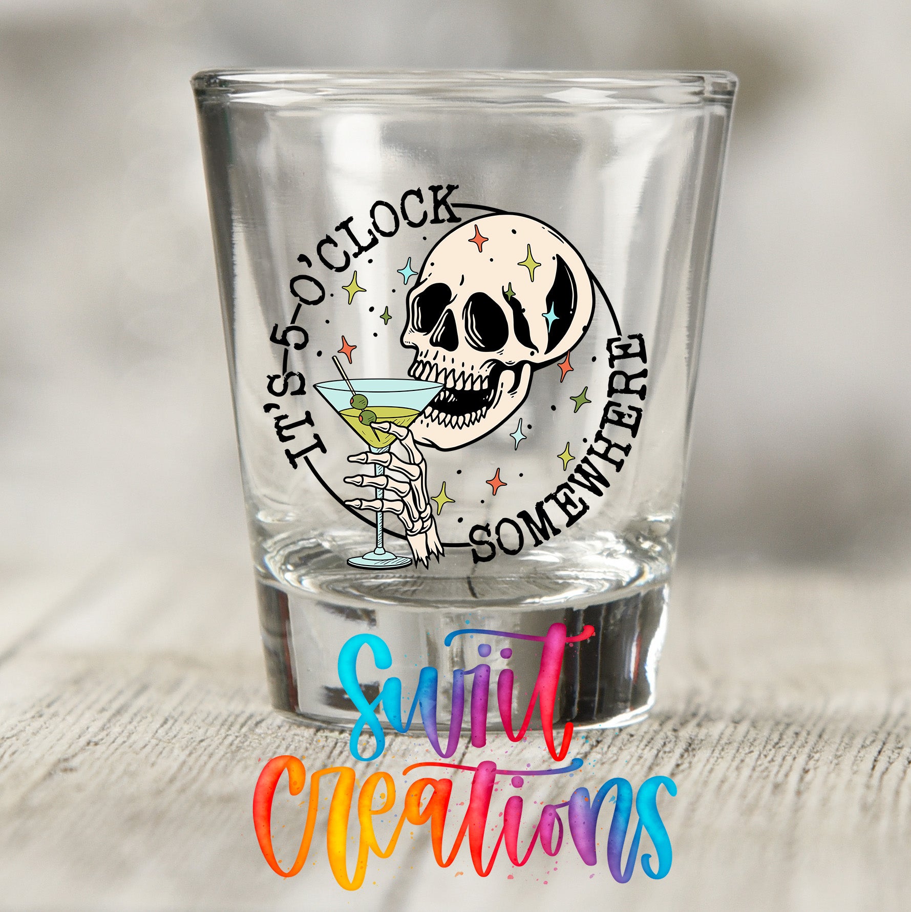 A shot glass with a skull and a cocktail glass inside, along with the text "It's 5 o'clock somewhere" and "Someway" written around the rim.