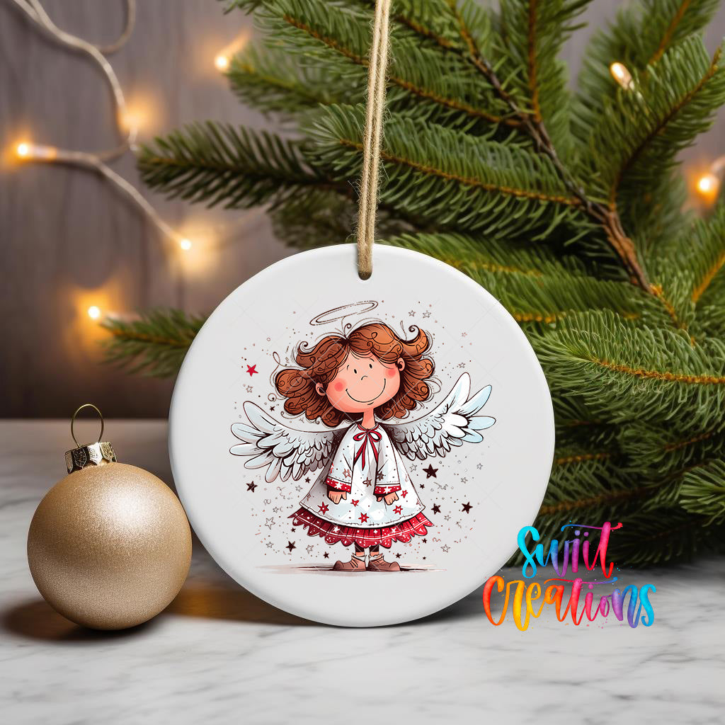 A white ceramic ornament featuring a smiling cartoon angel with wings, surrounded by stars, is displayed on a table next to a gold ornament and a Christmas tree.