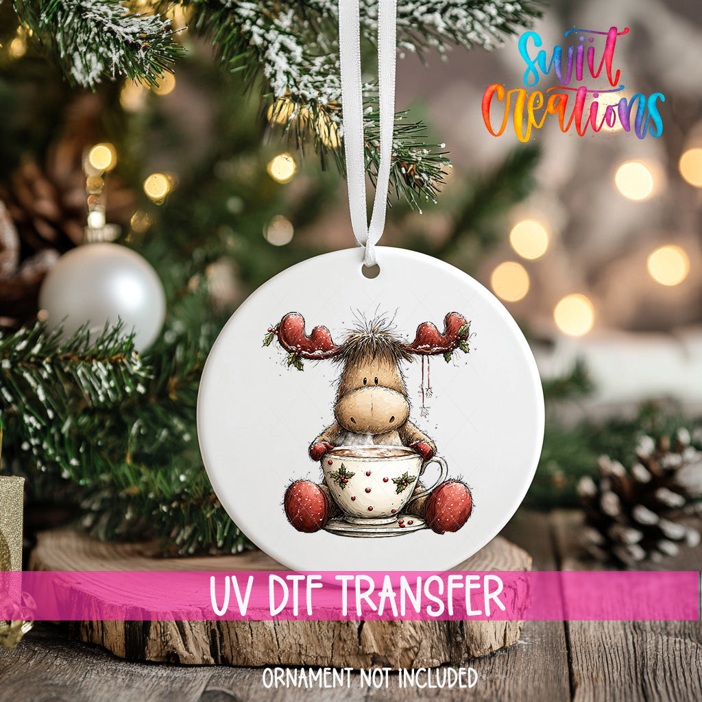 A white ornament with a cartoon moose sitting in a cup of hot chocolate is hanging on a Christmas tree.