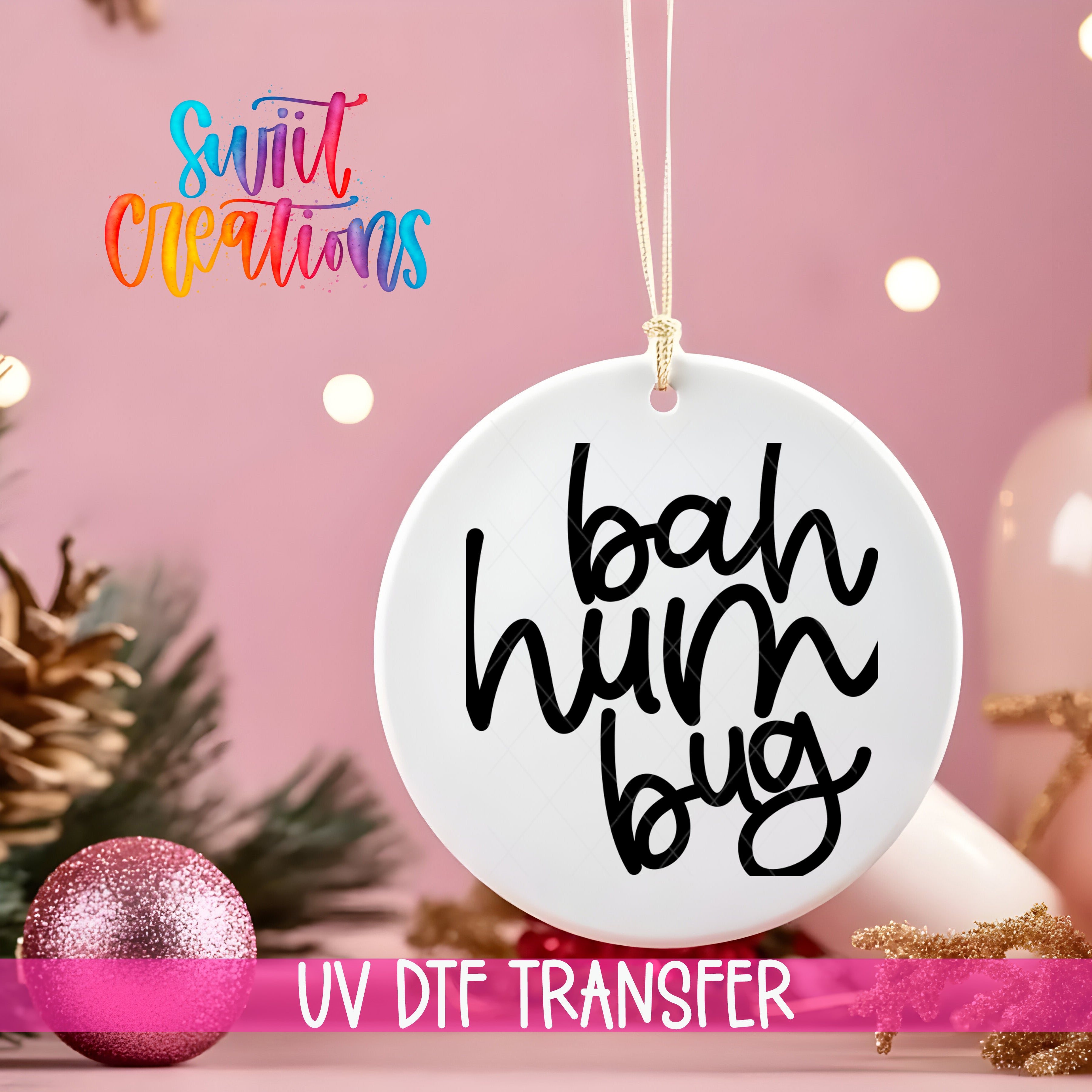 A white ornament with the text "bah hum bug" written on it, hanging against a pink background with Christmas decorations and a pink glitter ornament.