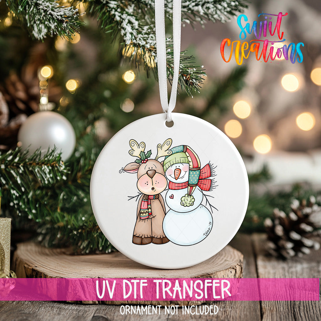 A white ceramic ornament with a deer and a snowman design hangs on a Christmas tree.