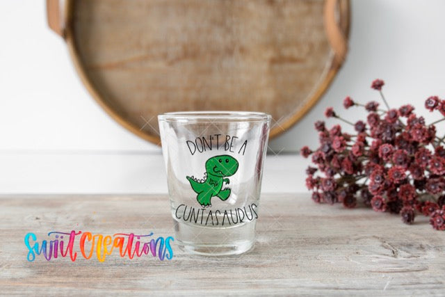 A shot glass with a cartoon dinosaur and the text "Don't be a Cuntasaurus" is placed on a wooden surface, with a bouquet of red flowers in the background.