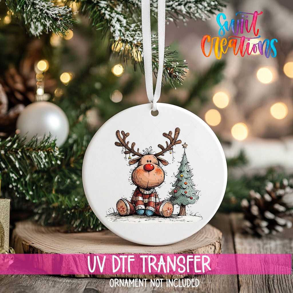 A white ornament with a cartoon image of a reindeer and a Christmas tree is hanging on a tree branch.