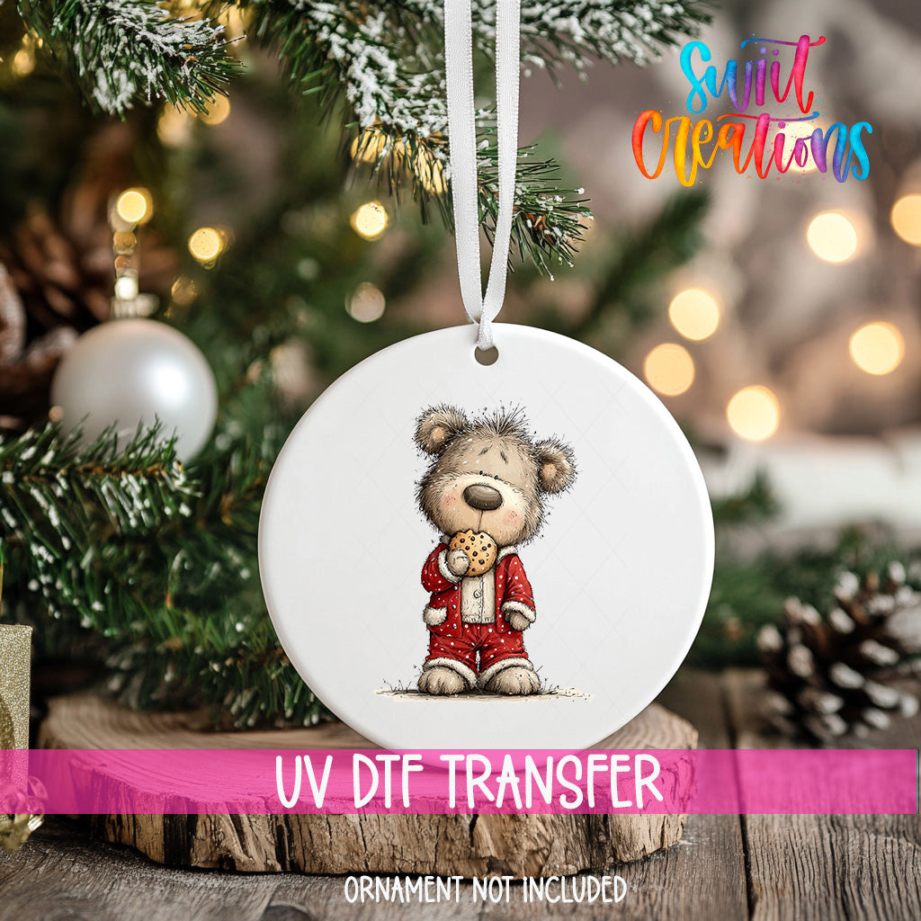 A white ornament with a teddy bear wearing a red outfit and holding a cookie is hanging on a Christmas tree.