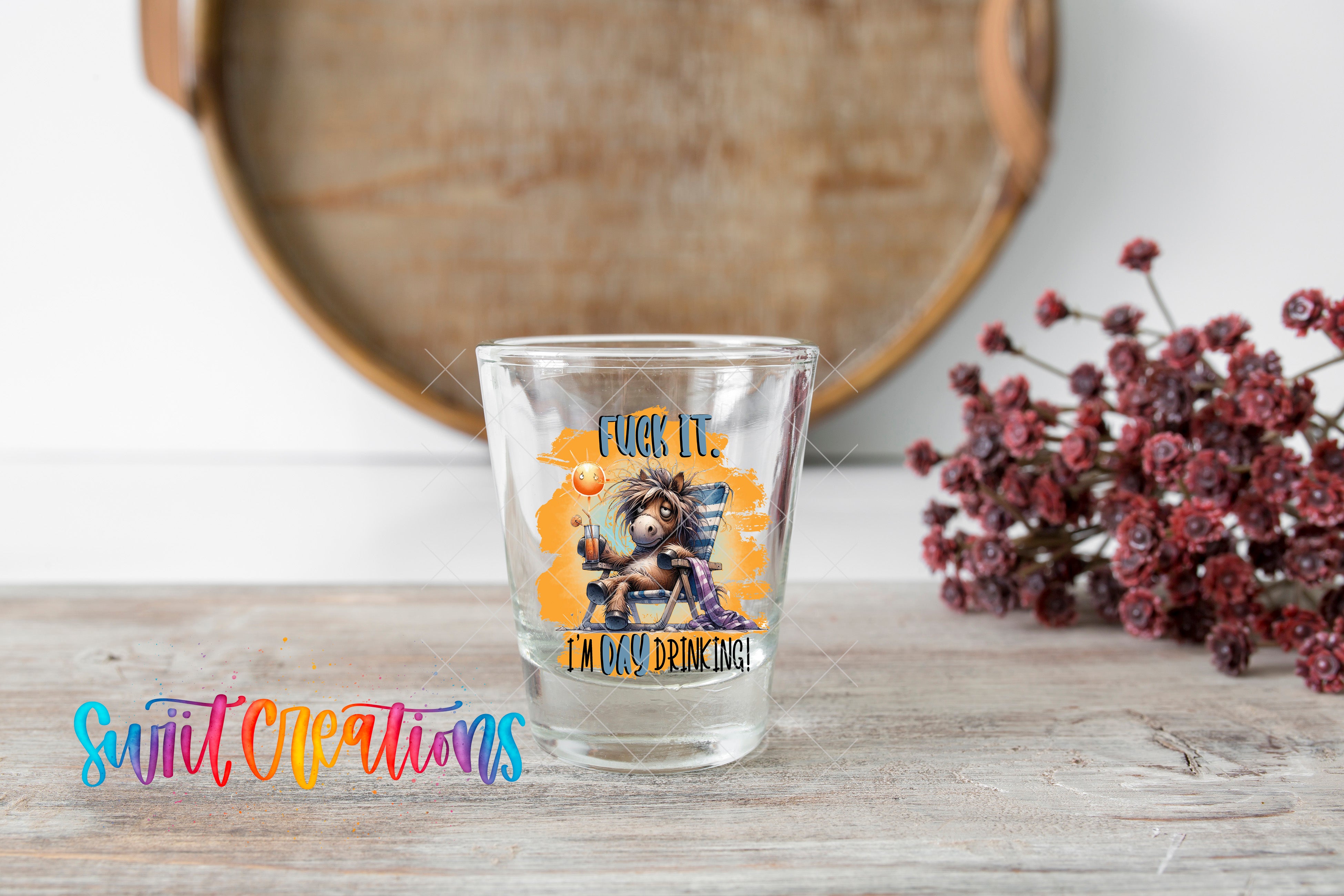 A shot glass with a colorful design and text on it, placed on a wooden surface next to a bunch of red flowers.