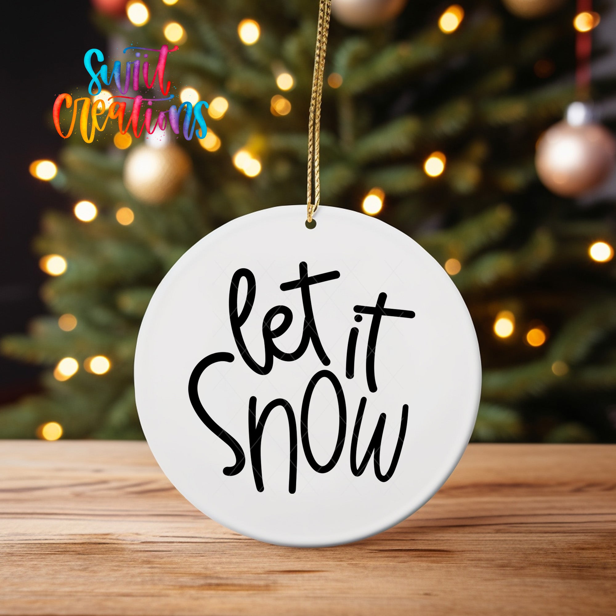 A white ornament with the text "let it snow" hanging from a string, placed on a wooden surface in front of a blurred Christmas tree with colorful lights.