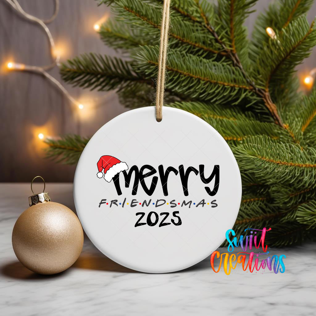 A white ceramic ornament with the text "MERRY FRIENDSMAS 2025" is displayed on a white marble surface, with a gold ornament and a Christmas tree in the background.
