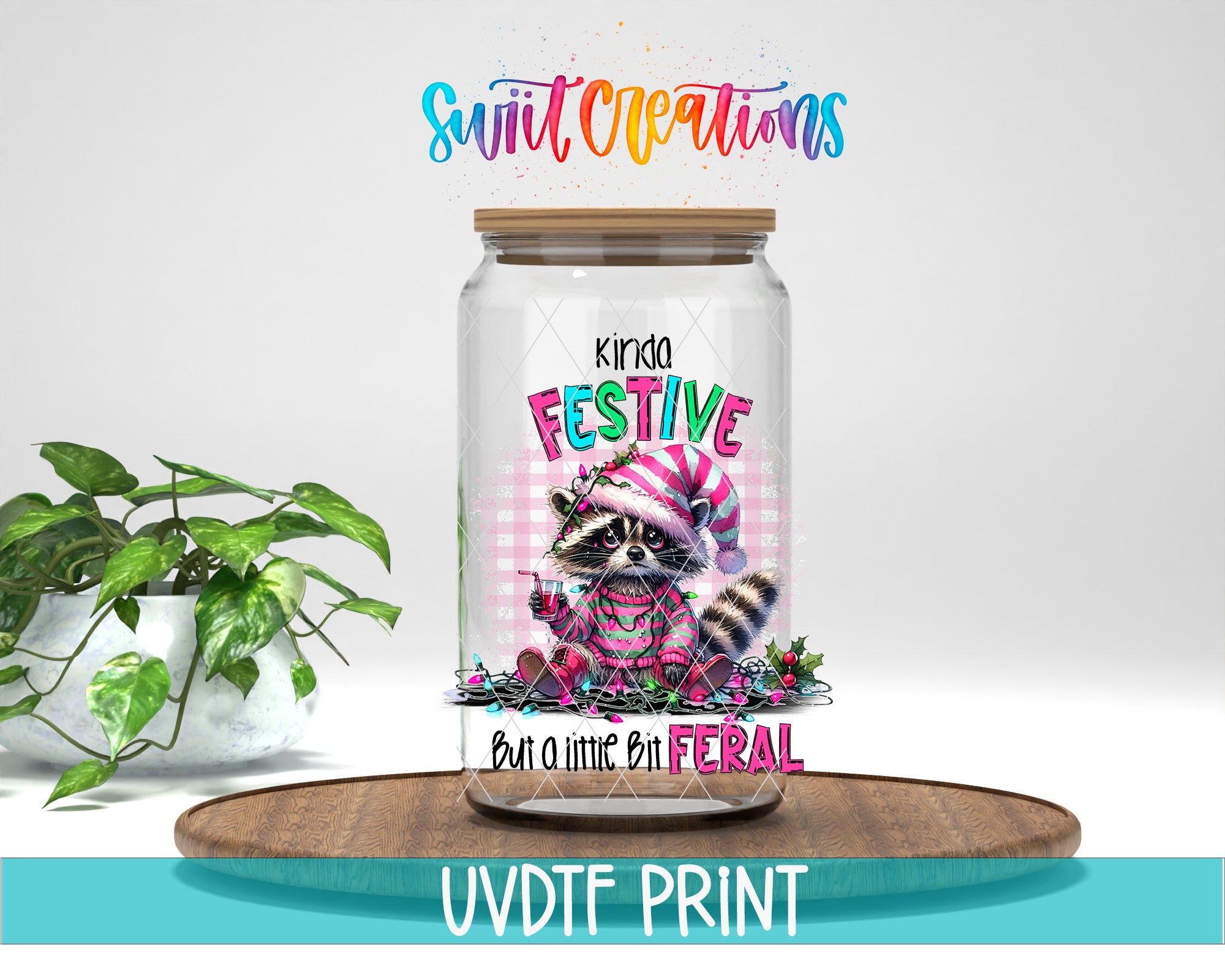 A jar with a raccoon wearing a festive hat and scarf, surrounded by holiday decorations, sits on a wooden tray. The jar is labeled "Kinda Festive" and "But a little bit Feral". The background is a plain white wall, and there is a potted plant to the left of the jar.