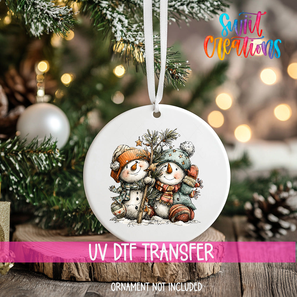 A white ornament with a snowman and pine tree design hangs on a Christmas tree, with a pink ribbon around it.