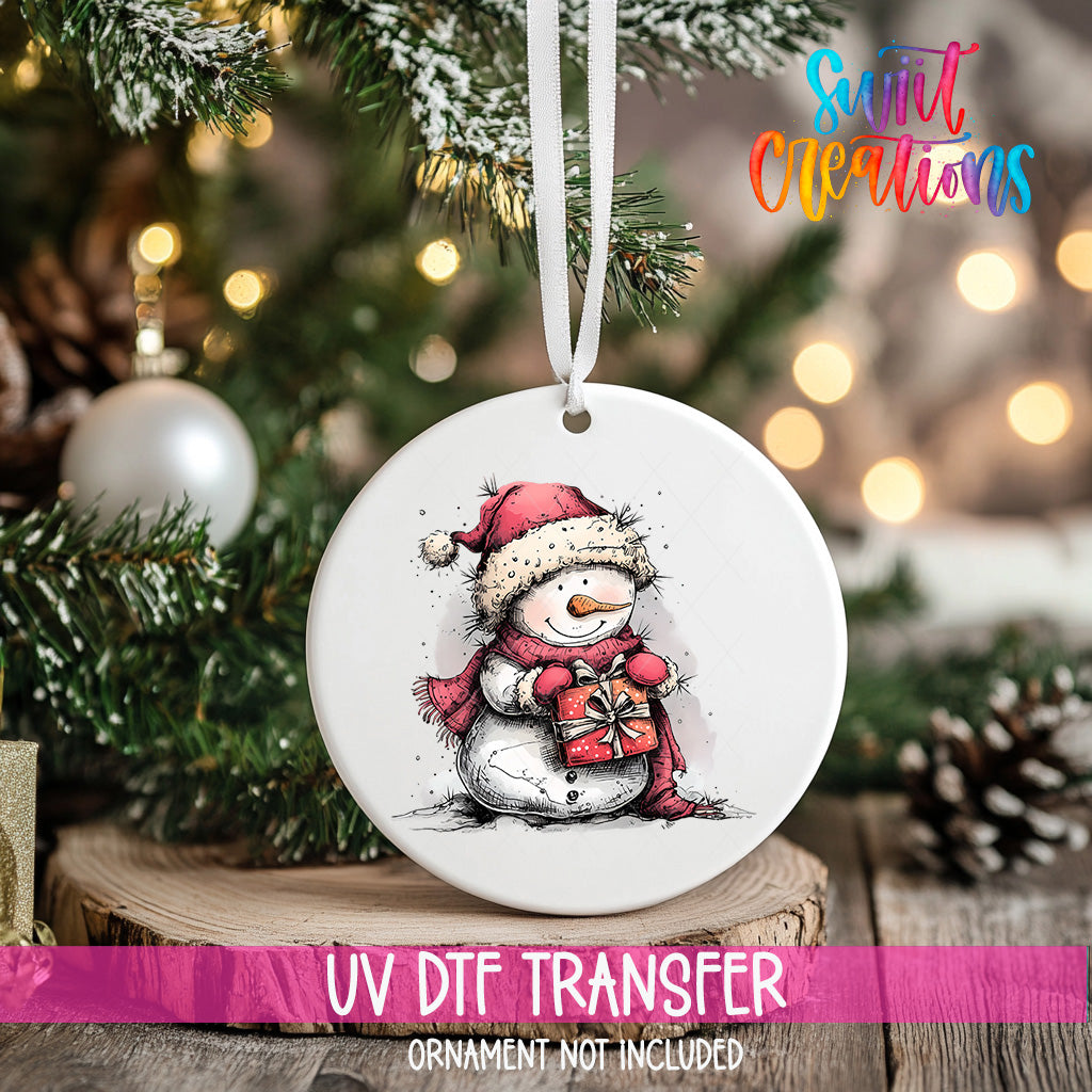 A white ceramic ornament with a snowman design is hanging on a Christmas tree, with a blurred background of pine branches and twinkling lights.