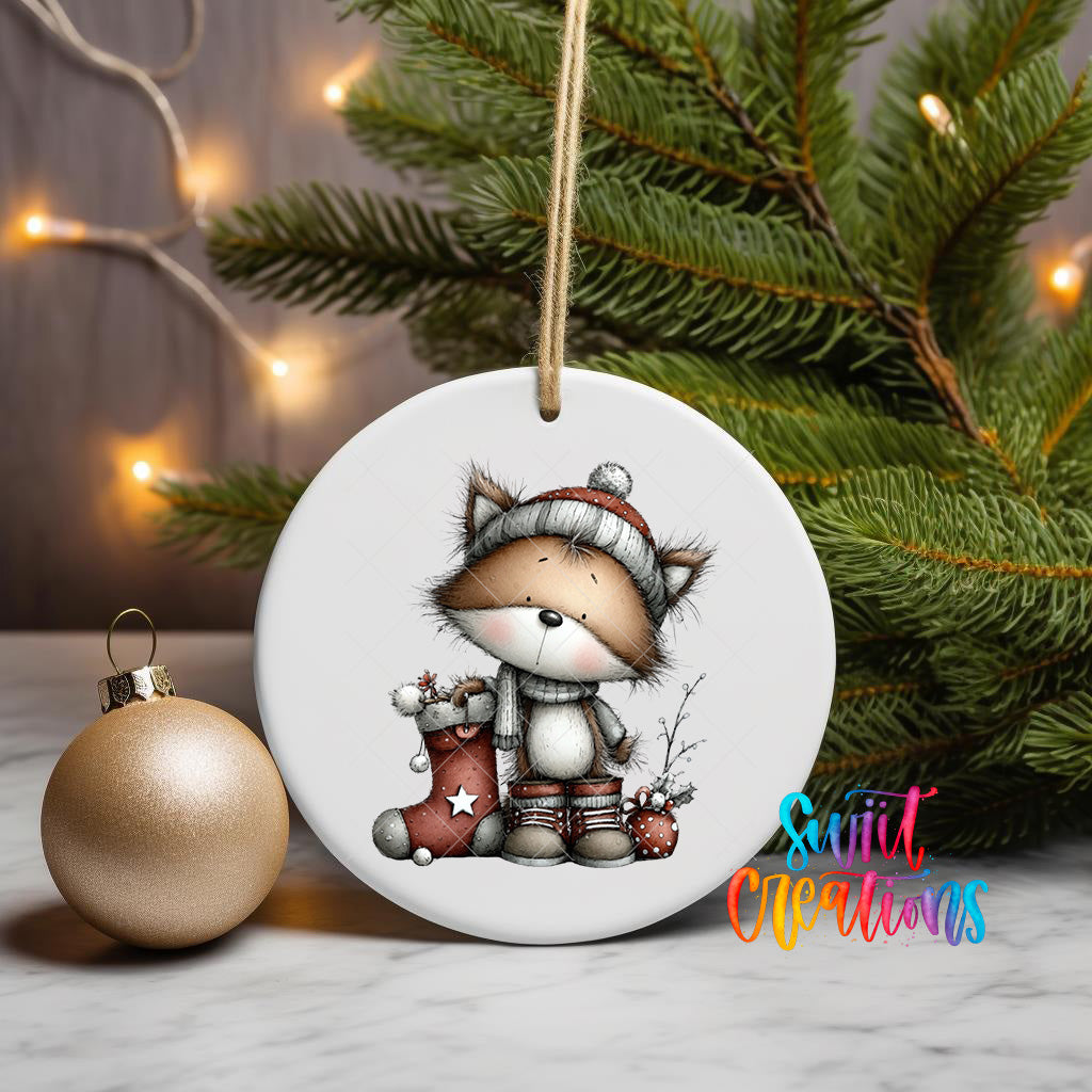 A white ceramic ornament featuring a cartoon fox wearing a red hat and holding a star-shaped ornament, placed on a marble surface next to a gold ornament and a Christmas tree with twinkling lights.