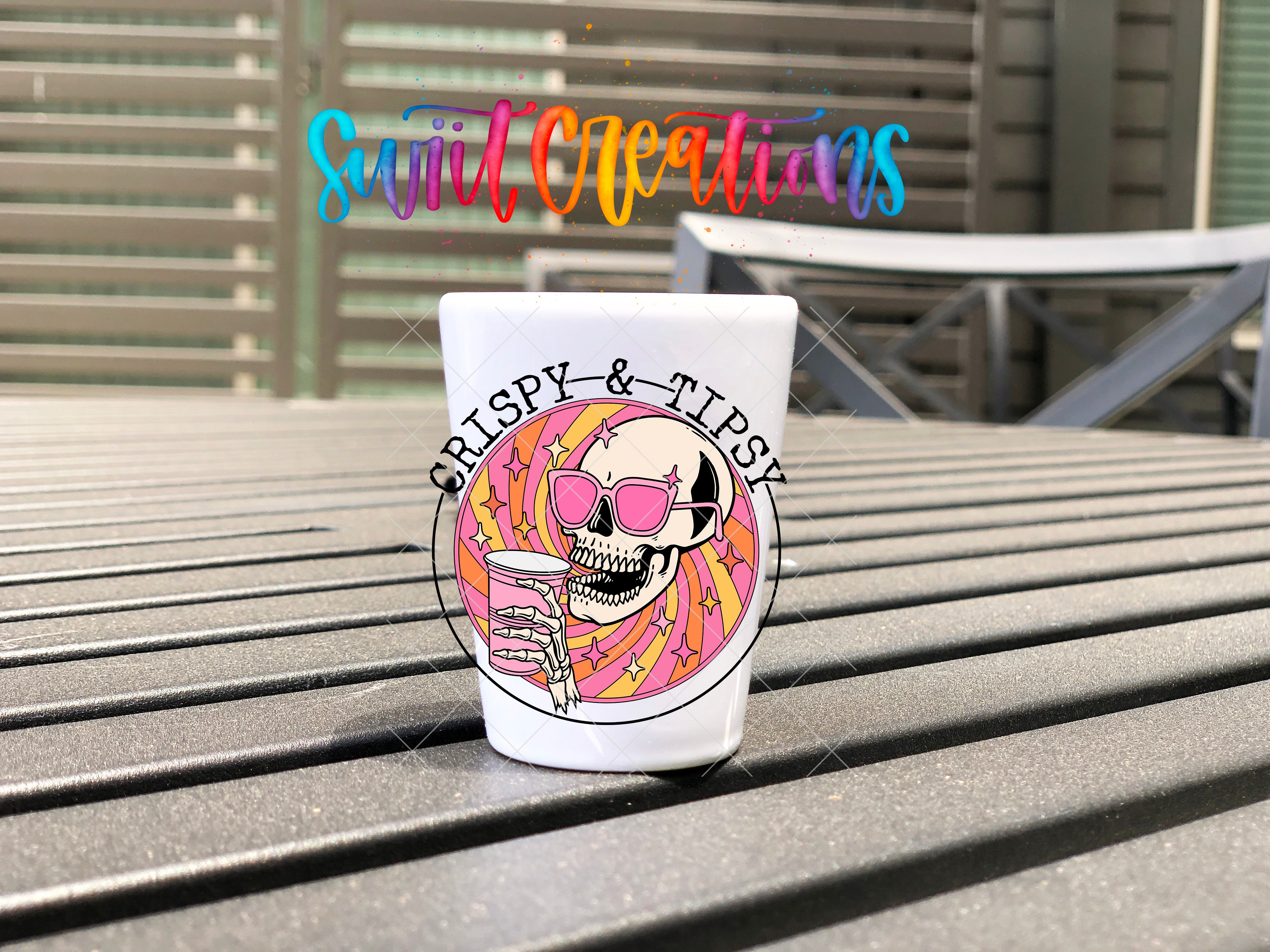 A white shot glass with a skull and sunglasses graphic, along with the text "CRISPY & TIPSY" printed on it, is placed on a wooden surface.